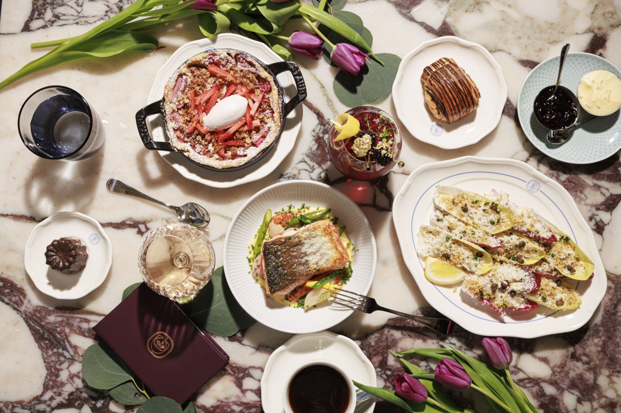 A marble table set with various dishes, including salad with grilled fish, baked pasta, stuffed endives, pastries, sauces, a drink, and tulip flowers scattered around, creating an elegant dining scene. captured by seattle's best food and hospitality photographer Brooke Fitts