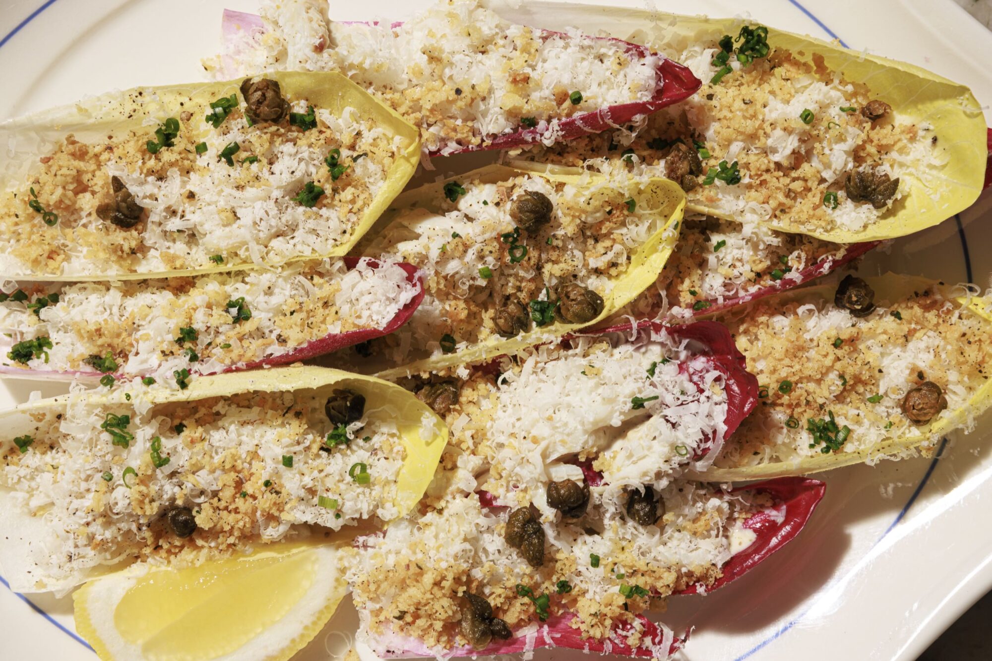 A plate of endive leaves topped with grated cheese, breadcrumbs, capers, and chopped herbs, served with lemon wedges on the side. The endive leaves are both yellow and purple. captured by seattle's best food and hospitality photographer Brooke Fitts