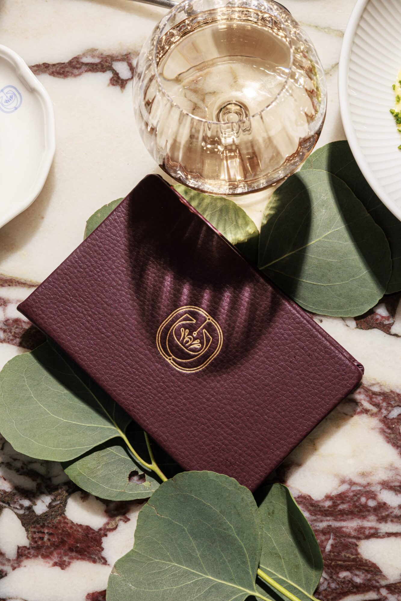 A burgundy textured menu with a gold emblem rests on green leaves atop a marble table, near a glass of rosé wine and a white plate. captured by seattle's best food and hospitality photographer Brooke Fitts
