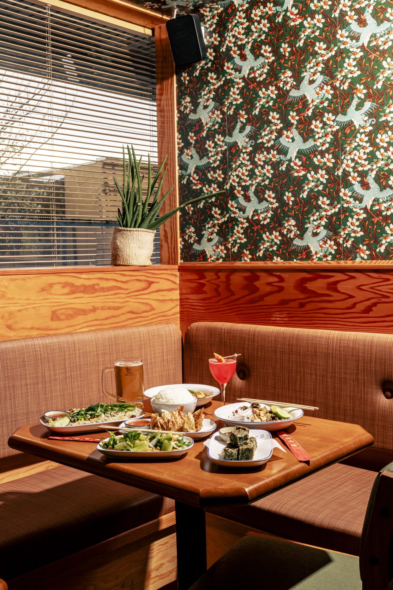 A cozy restaurant booth with a wooden table set with various dishes, drinks, and a potted plant by the window. Colorful floral wallpaper decorates the wall, with sunlight streaming through the blinds. captured by seattle's best food and hospitality photographer Brooke Fitts
