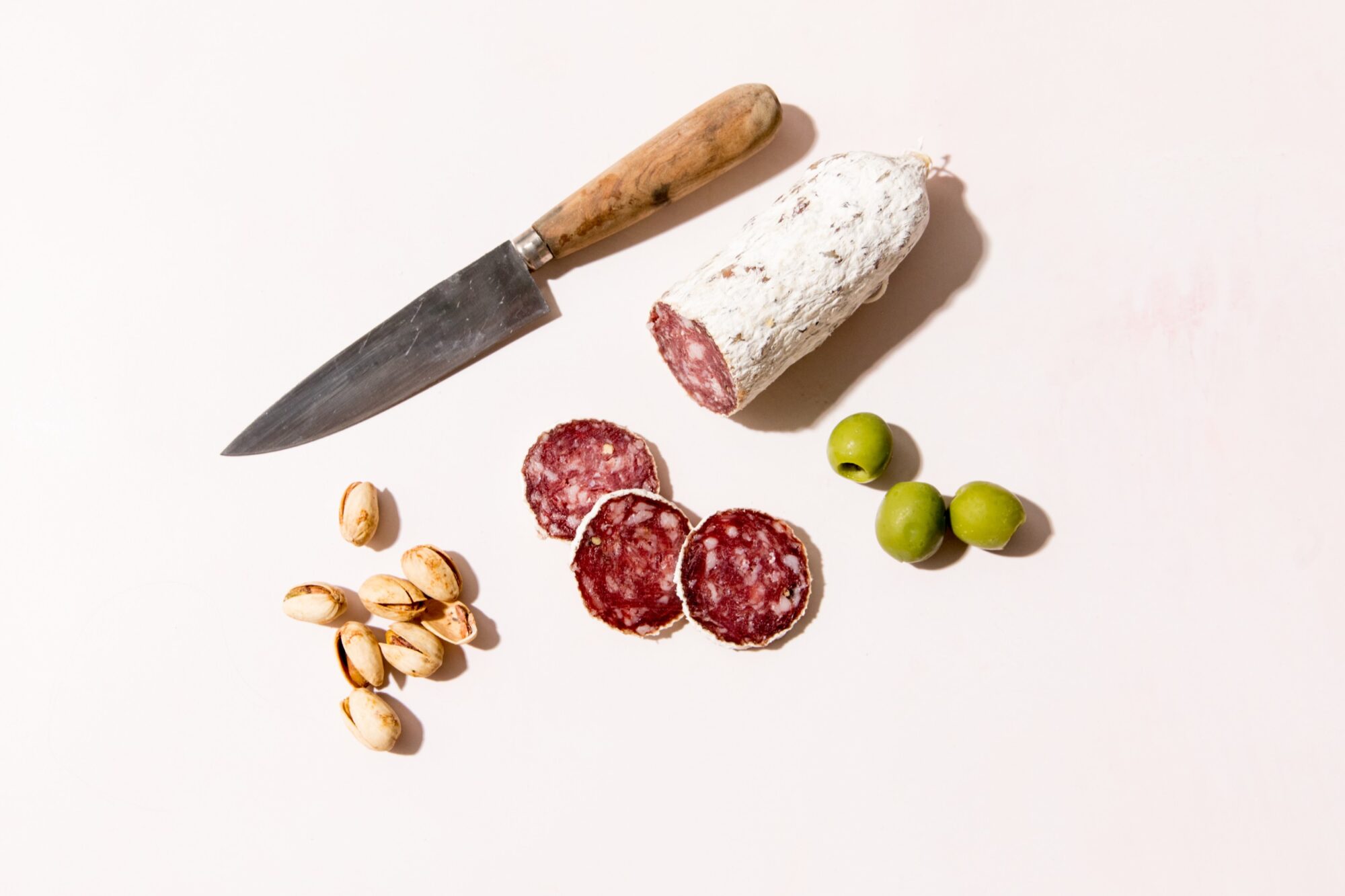 A knife, a partially sliced salami, three salami slices, a small pile of pistachios, and two green olives are arranged on a light-colored surface. captured by seattle's best food and hospitality photographer Brooke Fitts