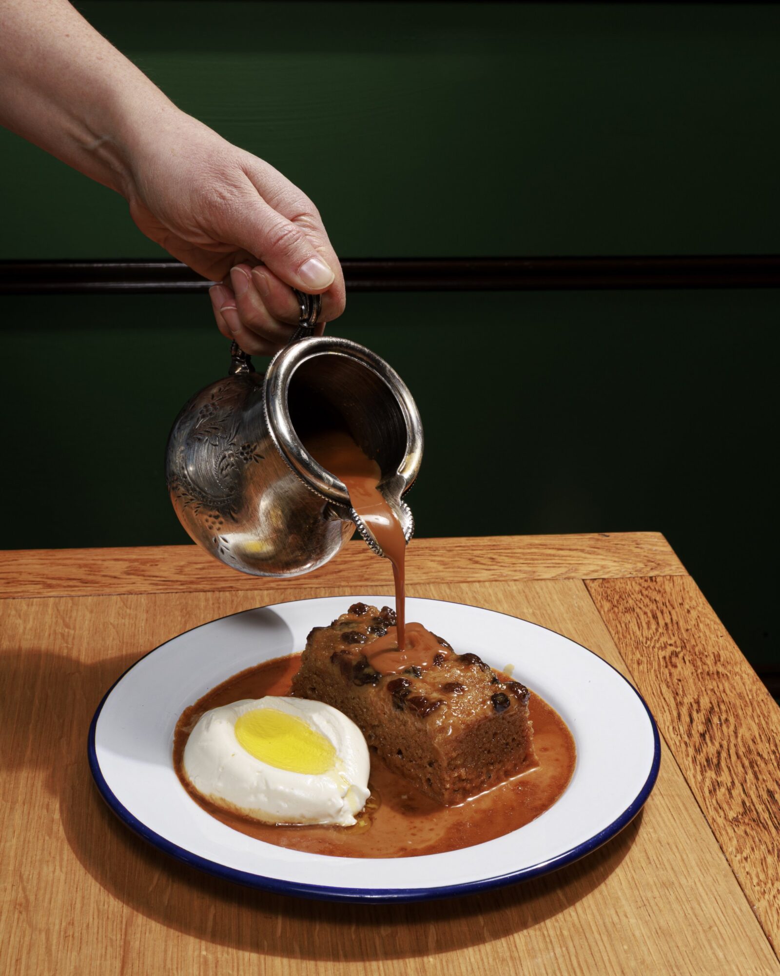 A hand pours brown sauce from a silver pitcher onto slices of bread pudding and a poached egg, all served on a white plate with a blue rim on a wooden table. captured by seattle's best food and hospitality photographer Brooke Fitts