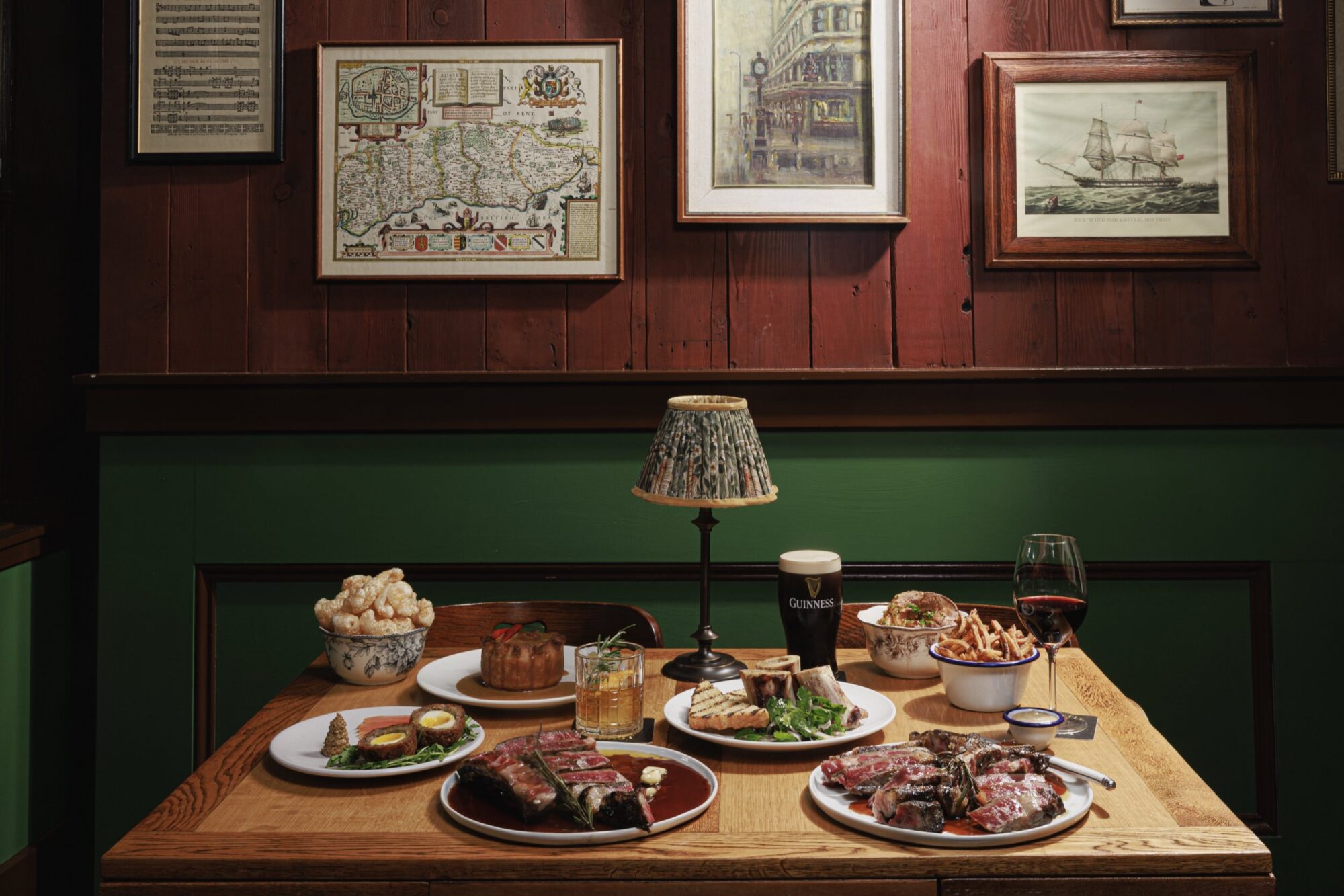A wooden table set with plates of ribs, steak, fries, bread, popcorn, and drinks including beer and wine, beneath a small lamp. The background features framed maps and artwork on wood-paneled walls. captured by seattle's best food and hospitality photographer Brooke Fitts