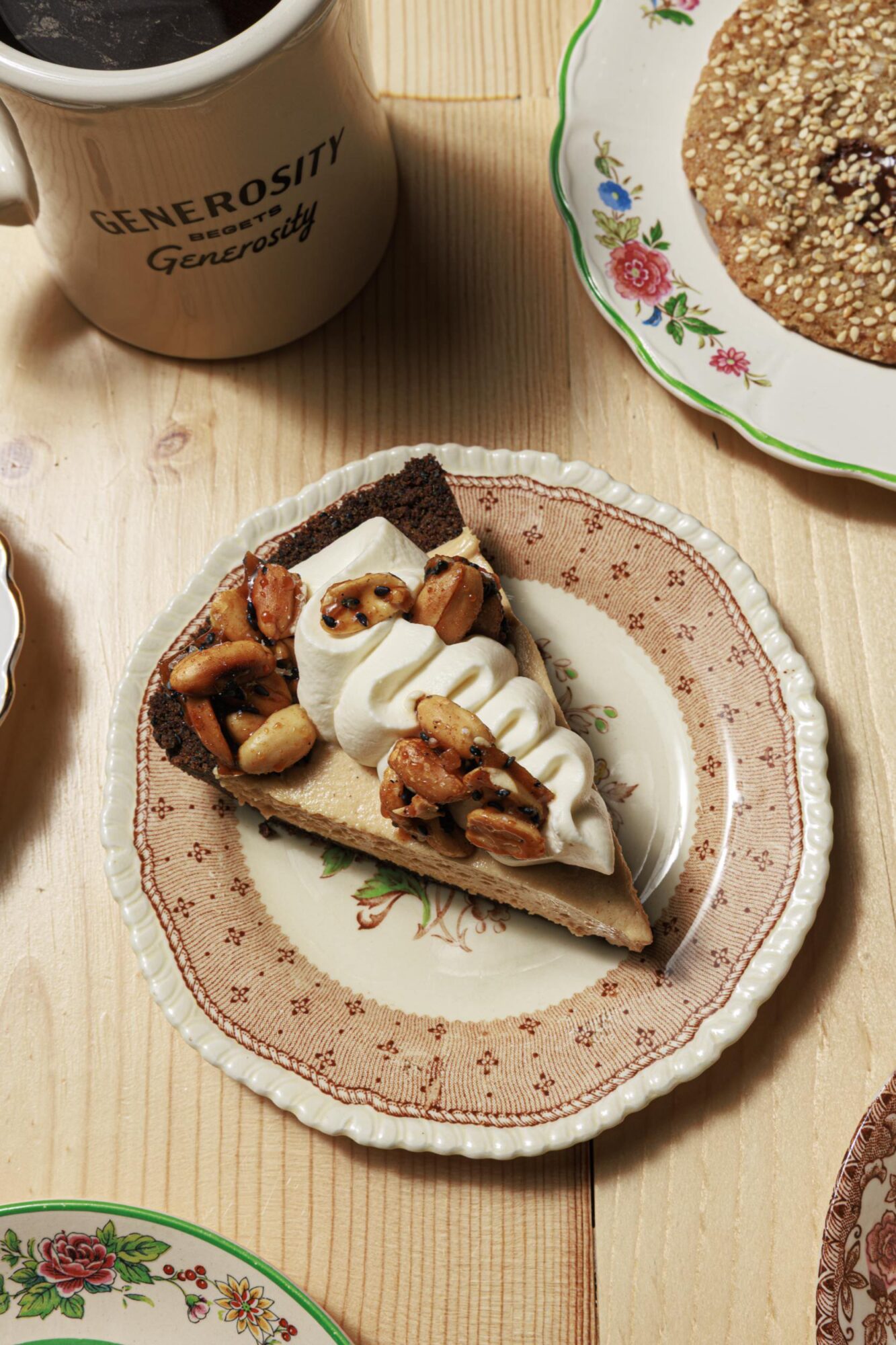 A slice of pie topped with whipped cream and nuts sits on a decorative plate. A mug labeled Generosity and a floral plate with a cookie are nearby on a light wooden table. captured by seattle's best food and hospitality photographer Brooke Fitts