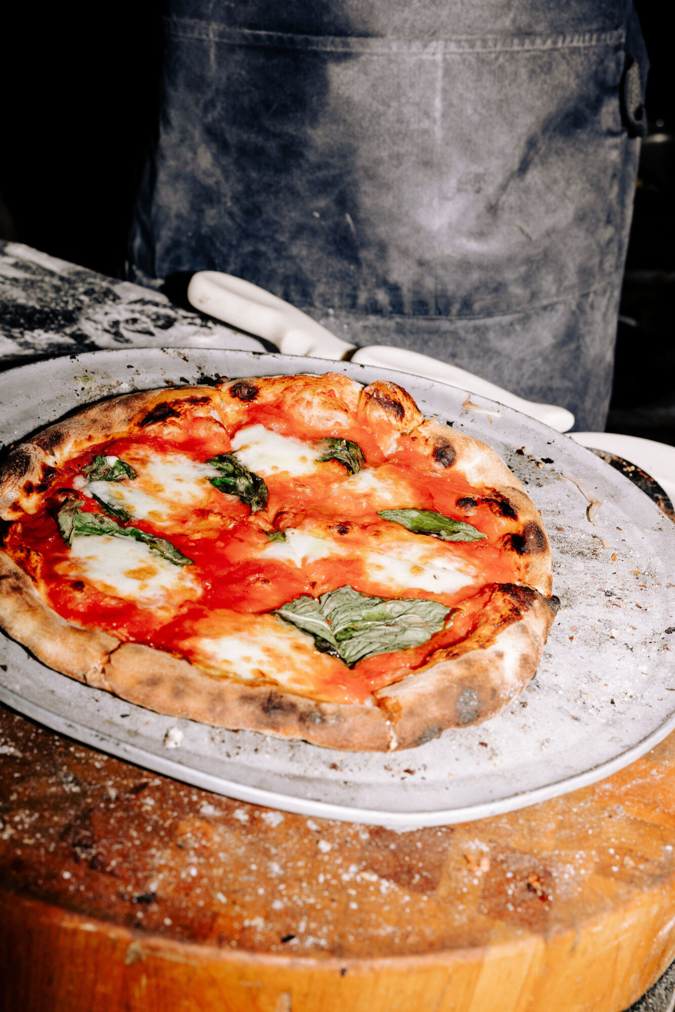 A freshly baked Margherita pizza with melted cheese, tomato sauce, and basil leaves sits on a metal tray atop a wooden surface. A person wearing a gray apron stands behind the pizza. captured by seattle's best food and hospitality photographer Brooke Fitts