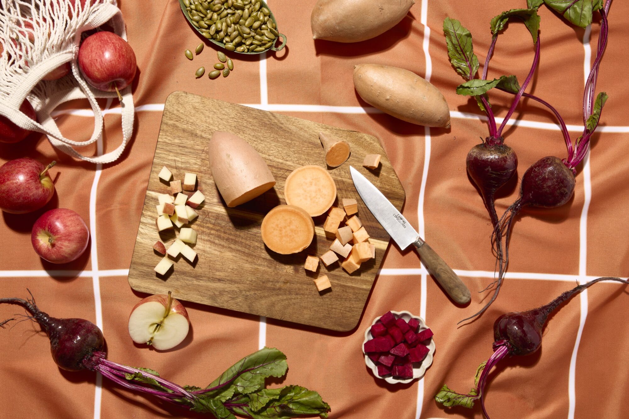 A wooden cutting board with a knife, diced and whole sweet potatoes, diced beets in a bowl, whole apples, halved apple, beets with leaves, and pumpkin seeds on an orange checked cloth. captured by seattle's best food and hospitality photographer Brooke Fitts