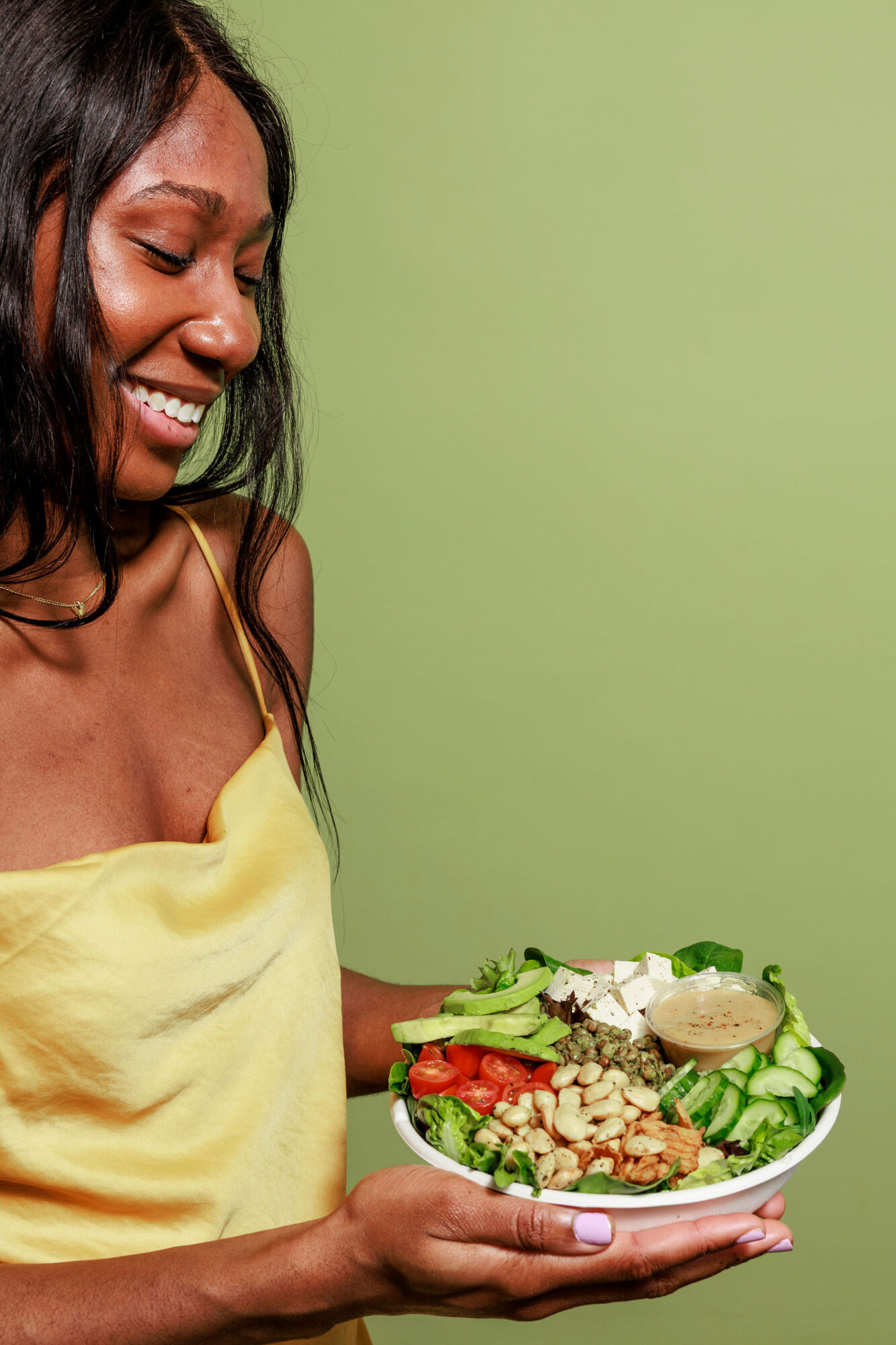 A smiling woman in a yellow dress holds a colorful salad bowl filled with greens, tofu, tomatoes, cucumbers, nuts, seeds, and a dollop of dressing, against a green background. captured by seattle's best food and hospitality photographer Brooke Fitts