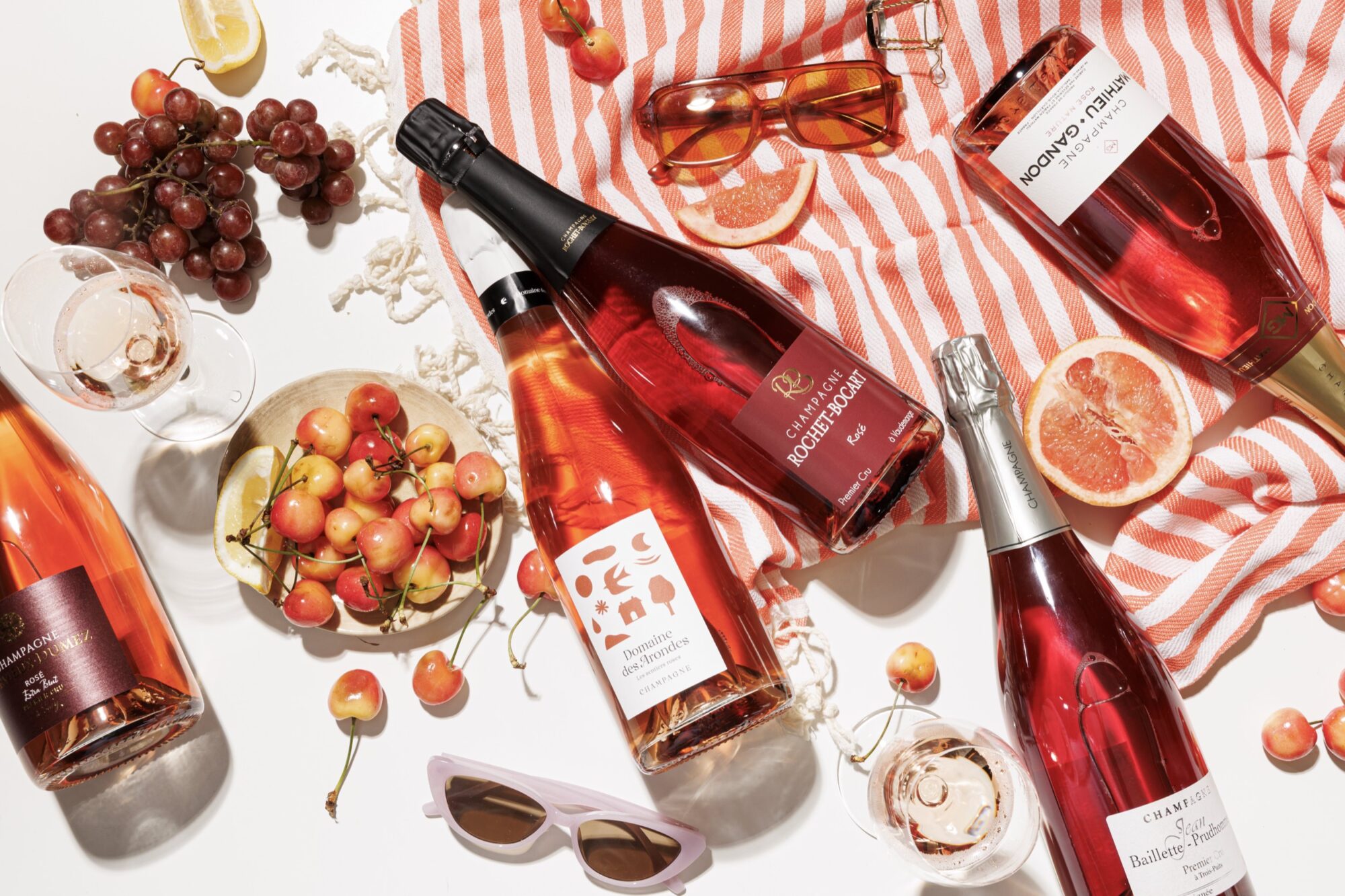 Bottles of rosé wine, glasses, cherries, grapes, grapefruit slices, and sunglasses are arranged on a white surface with a red-striped cloth, creating a bright, summery picnic scene. captured by seattle's best food and hospitality photographer Brooke Fitts