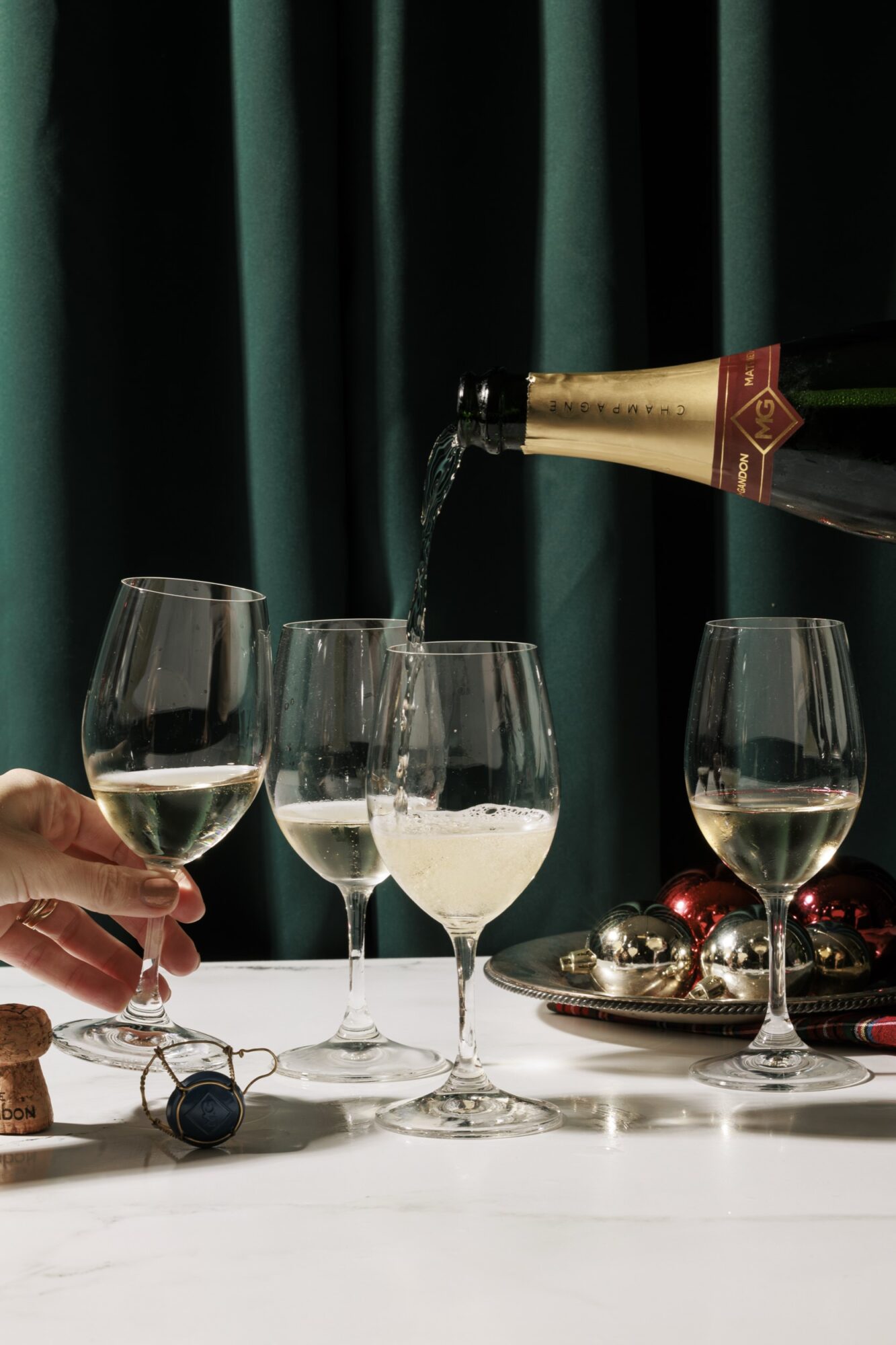 A bottle of sparkling wine is being poured into one of three wine glasses on a table. A hand holds one glass, and a plate with ornaments sits nearby. Green curtains hang in the background. captured by seattle's best food and hospitality photographer Brooke Fitts