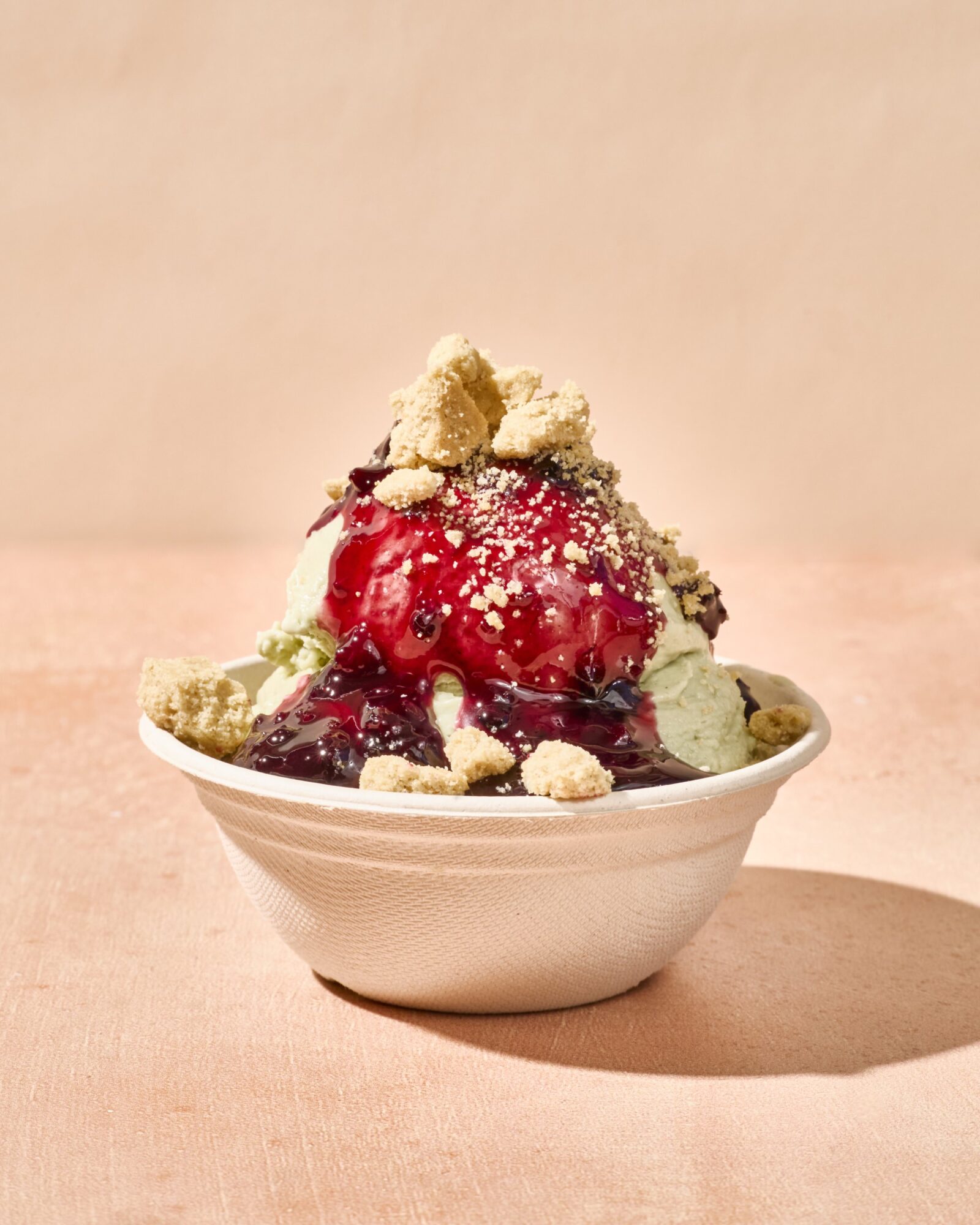 A bowl of ice cream topped with berry sauce and crumbled cookies, set against a light peach background. captured by seattle's best food and hospitality photographer Brooke Fitts