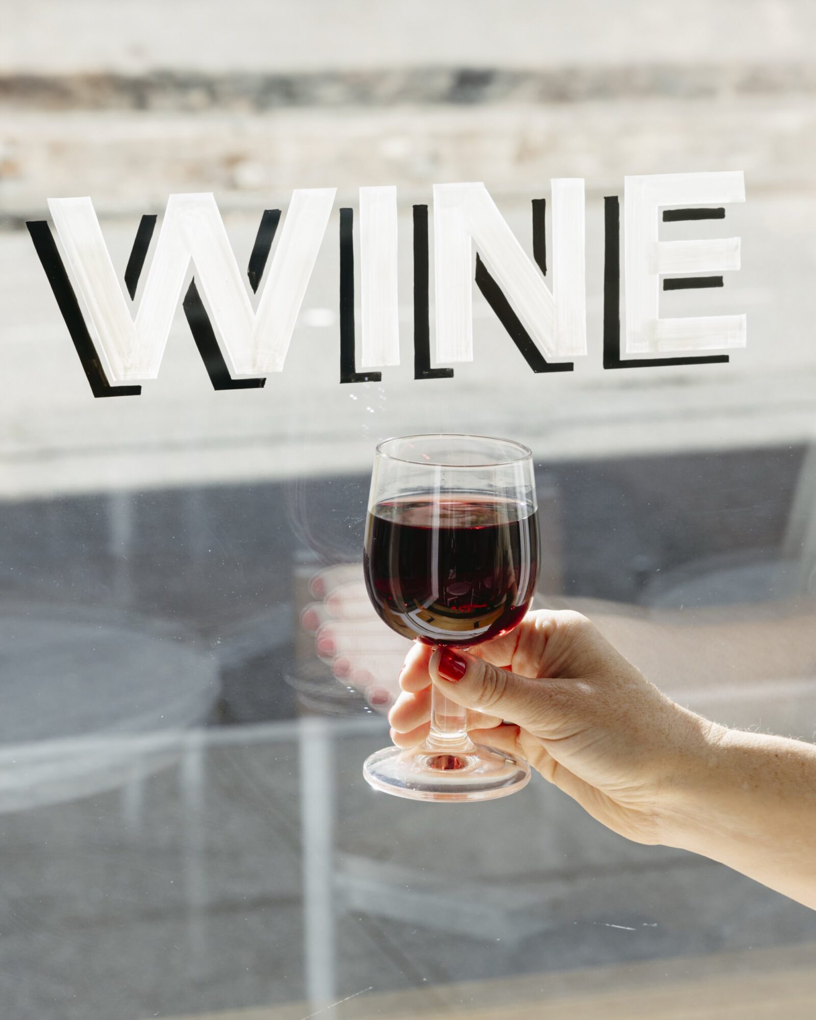 A hand holding a glass of red wine in front of a window with the word WINE written in large white letters on the glass. captured by seattle's best food and hospitality photographer Brooke Fitts