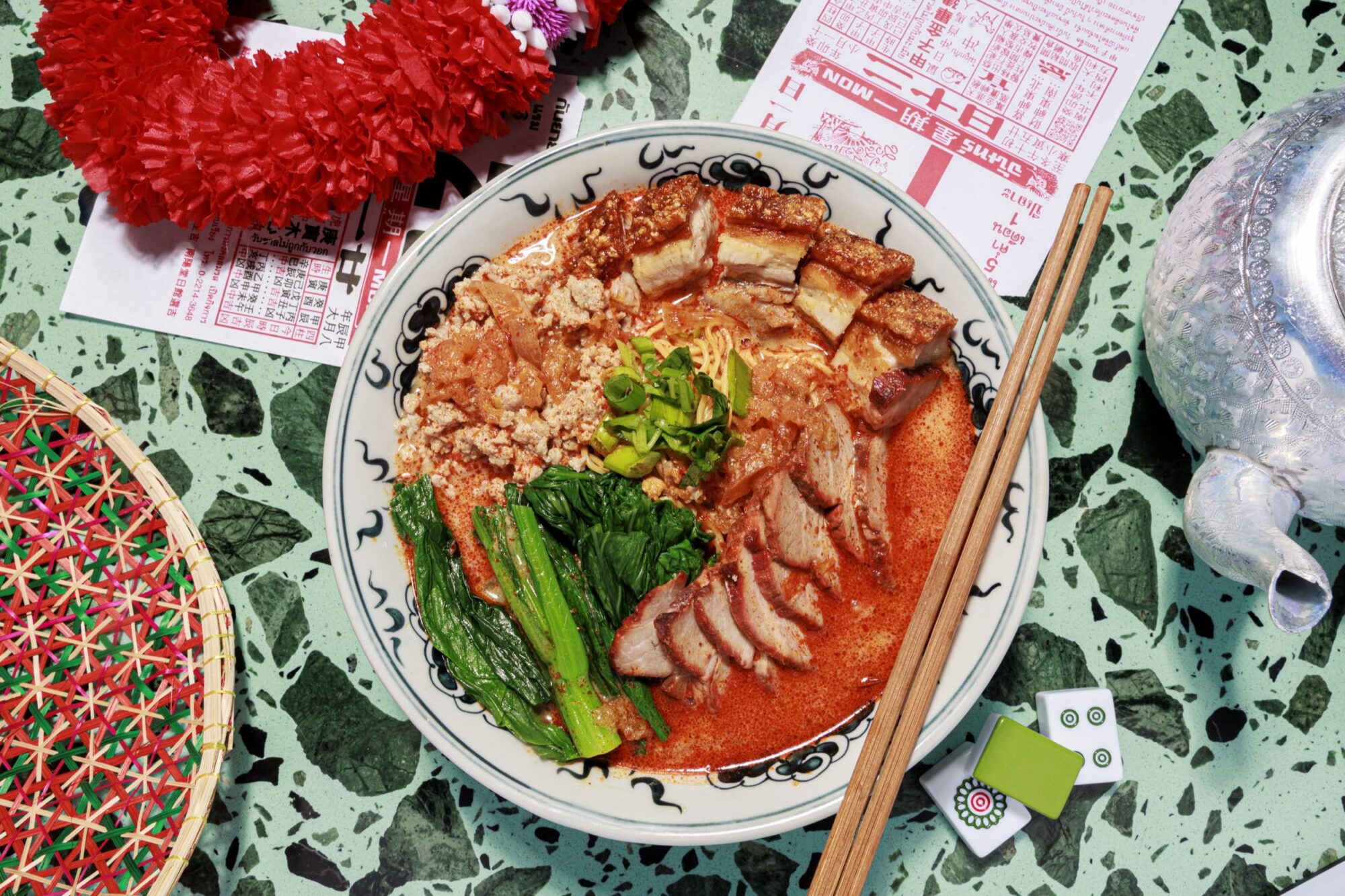 A bowl of vibrant noodle soup with sliced pork, crispy pork belly, greens, and scallions in red broth, served with chopsticks on a patterned table surrounded by festive items and game tiles. captured by seattle's best food and hospitality photographer Brooke Fitts