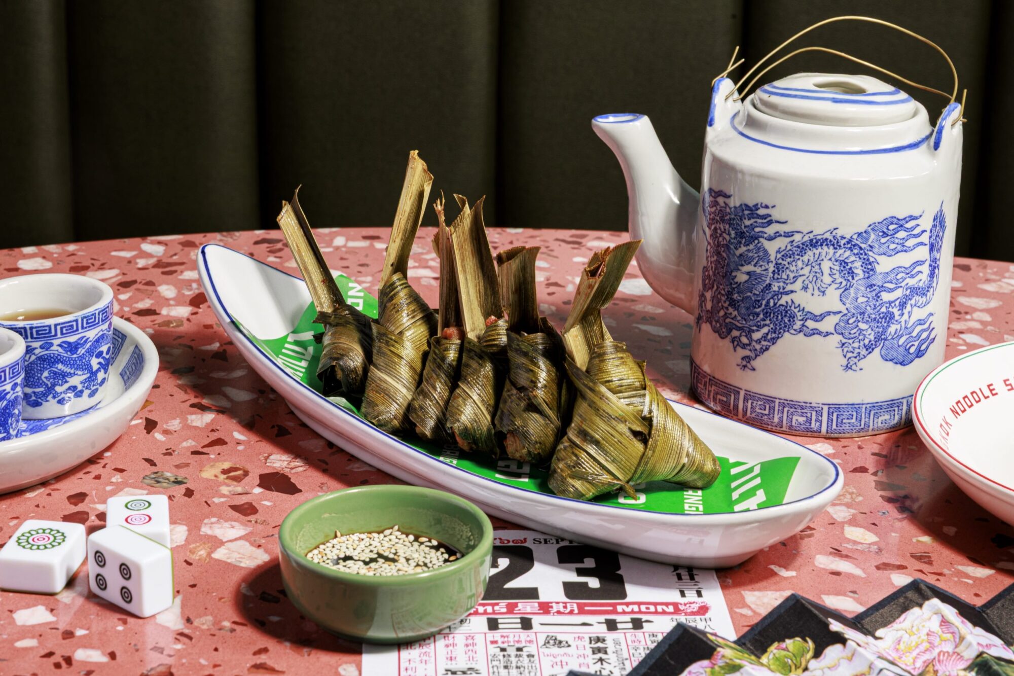 A plate of pandan leaf-wrapped dumplings sits on a terrazzo table beside a blue and white teapot, blue-patterned teacups, a small bowl of sesame seeds, Mahjong tiles, and paper slips. captured by seattle's best food and hospitality photographer Brooke Fitts