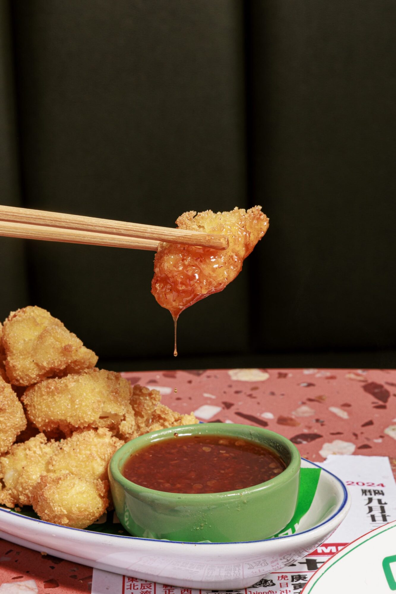 A pair of chopsticks holds a piece of crispy fried food dipped in sauce, with more pieces stacked on a plate beside a small green bowl of reddish dipping sauce on a pink speckled table. captured by seattle's best food and hospitality photographer Brooke Fitts