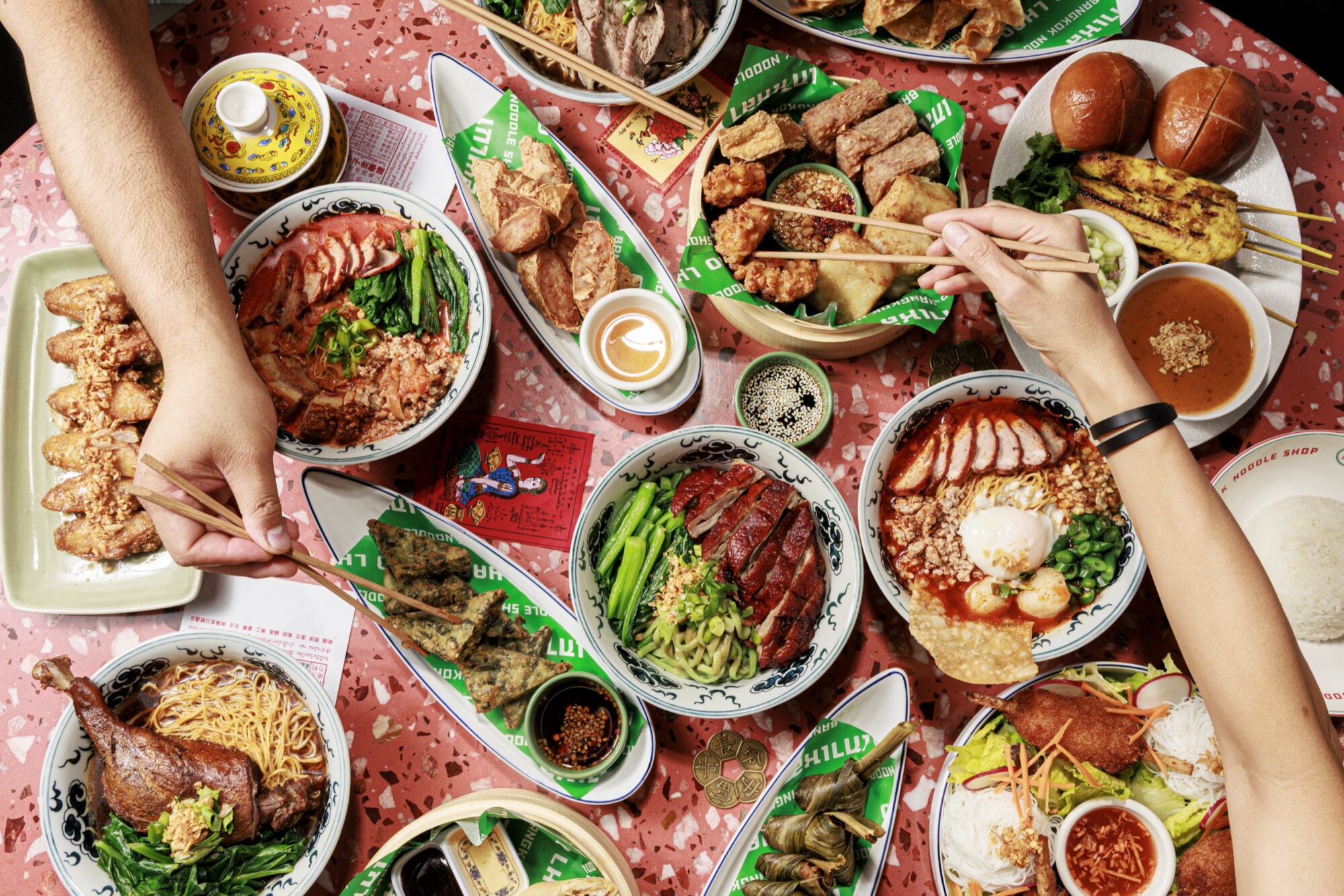 A table filled with colorful Asian dishes, including noodles, rice bowls with roasted meats, dumplings, fried chicken, greens, and dipping sauces. Two people use chopsticks to pick food from the shared plates. captured by seattle's best food and hospitality photographer Brooke Fitts
