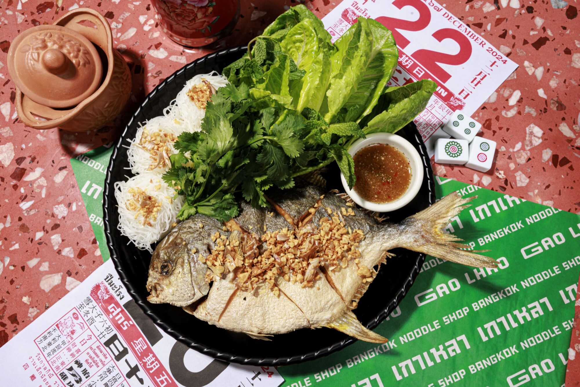 A plate with a whole fried fish topped with fried garlic, served with rice noodles, fresh herbs, lettuce, and dipping sauce. The dish sits on colorful papers and a terrazzo surface with a teapot and mahjong tile nearby. captured by seattle's best food and hospitality photographer Brooke Fitts