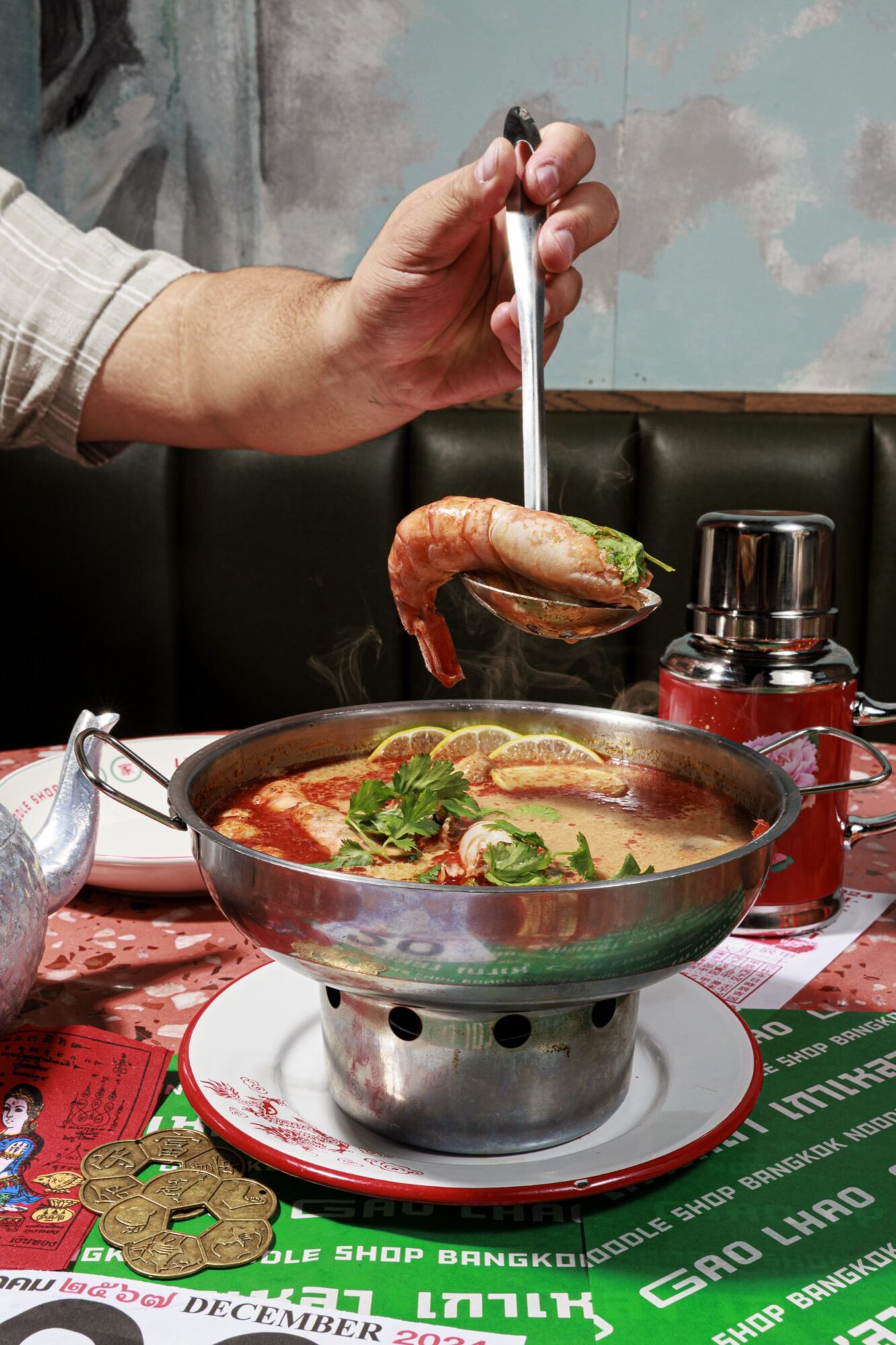 A hand holds a ladle scooping shrimp, herbs, and broth from a metal hot pot filled with tom yum soup, set on a colorful, decorated table at a restaurant. captured by seattle's best food and hospitality photographer Brooke Fitts