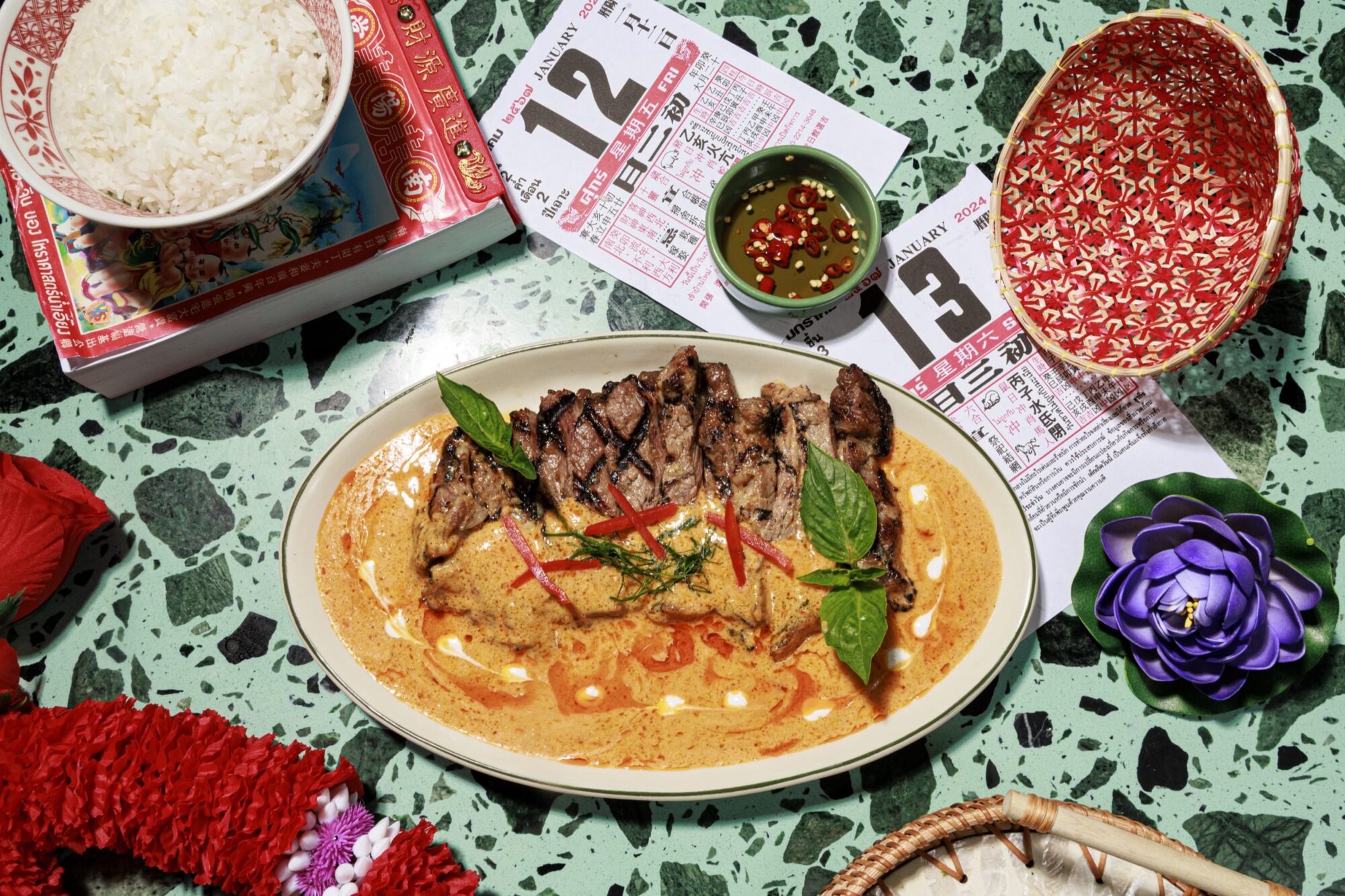 Grilled meat with red curry sauce garnished with herbs, served on an oval plate, sits on a green speckled table with rice, sauce, a purple flower, lottery tickets, and a woven basket nearby. captured by seattle's best food and hospitality photographer Brooke Fitts