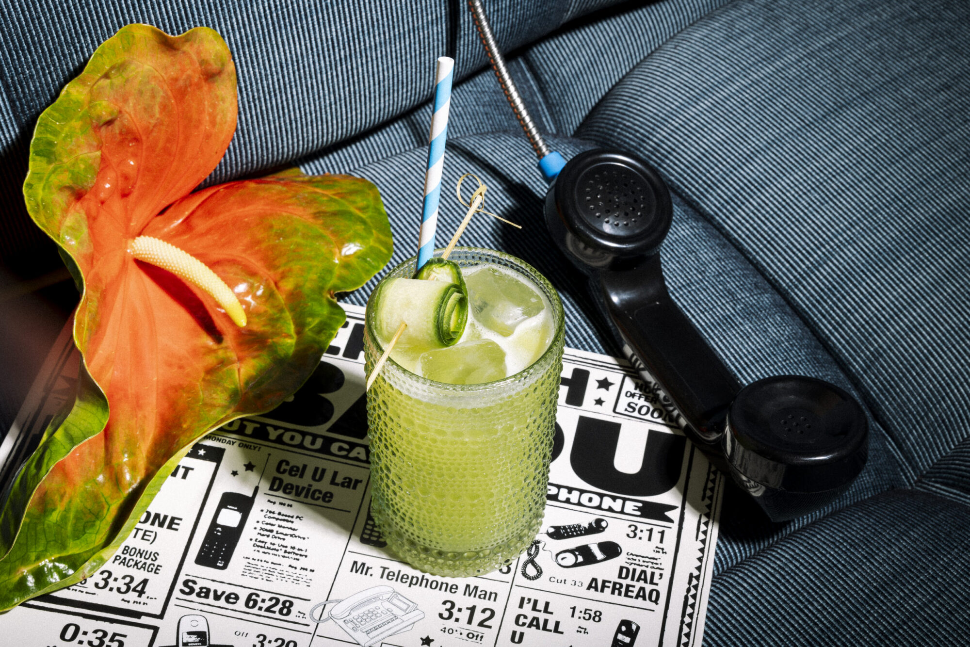 A green cocktail with cucumber garnish and a striped straw sits on a retro magazine next to an orange anthurium flower and a black rotary phone handset, all on a blue upholstered chair. captured by seattle's best food and hospitality photographer Brooke Fitts