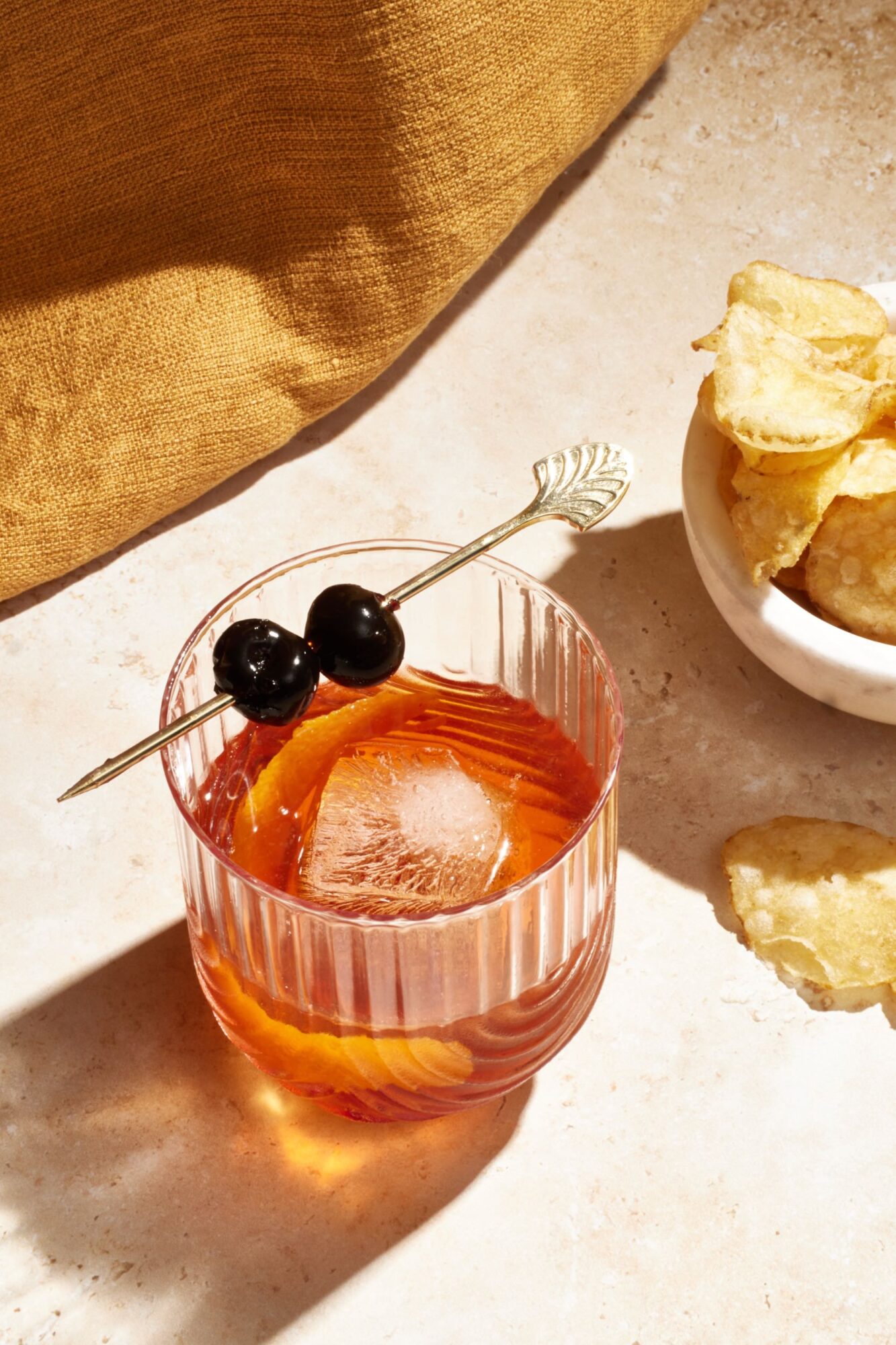 A glass of amber-colored cocktail with a large ice cube and two dark cherries on a decorative skewer, placed beside a small bowl of potato chips on a light stone surface. captured by seattle's best food and hospitality photographer Brooke Fitts
