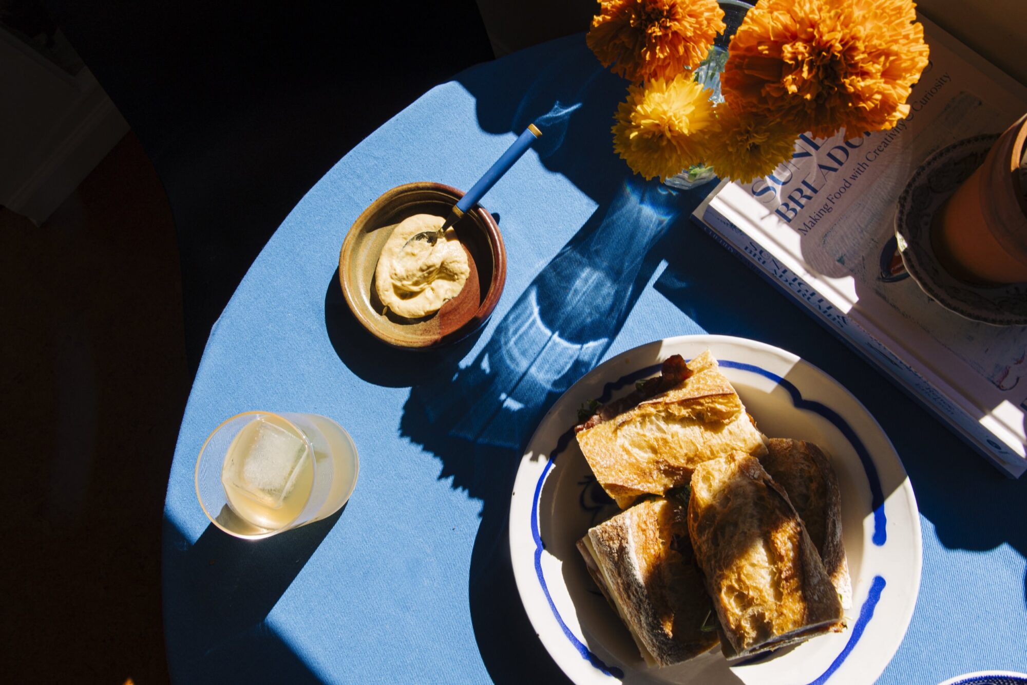 A blue tablecloth with a plate of toasted sandwiches, a bowl of dip with a spoon, a glass with a large ice cube, and a vase of orange flowers in sunlight. Books and a potted plant are partially visible on the side. captured by seattle's best food and hospitality photographer Brooke Fitts