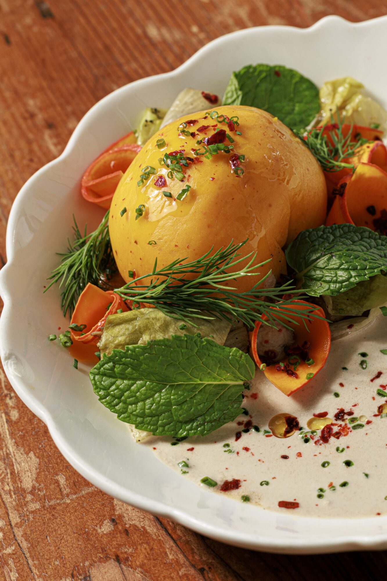 A colorful dish featuring a yellow tomato, fresh mint and dill leaves, carrot ribbons, lettuce, and creamy white sauce, garnished with herbs and red pepper flakes, served on a white plate over a wooden table. captured by seattle's best food and hospitality photographer Brooke Fitts