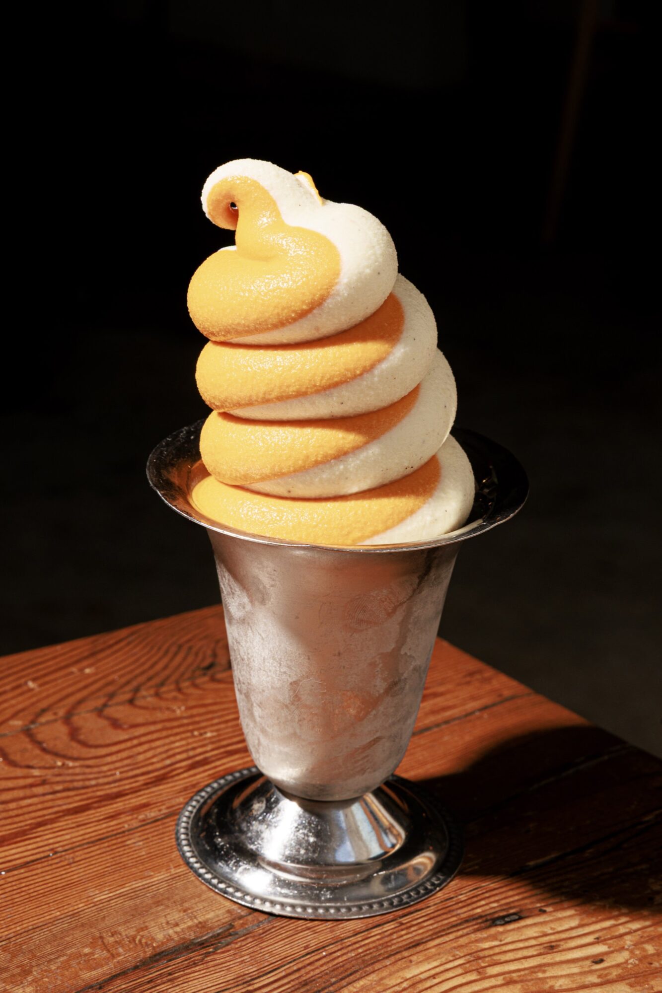 A metal cup filled with swirled orange and vanilla soft-serve ice cream sits on a wooden surface against a dark background. captured by seattle's best food and hospitality photographer Brooke Fitts