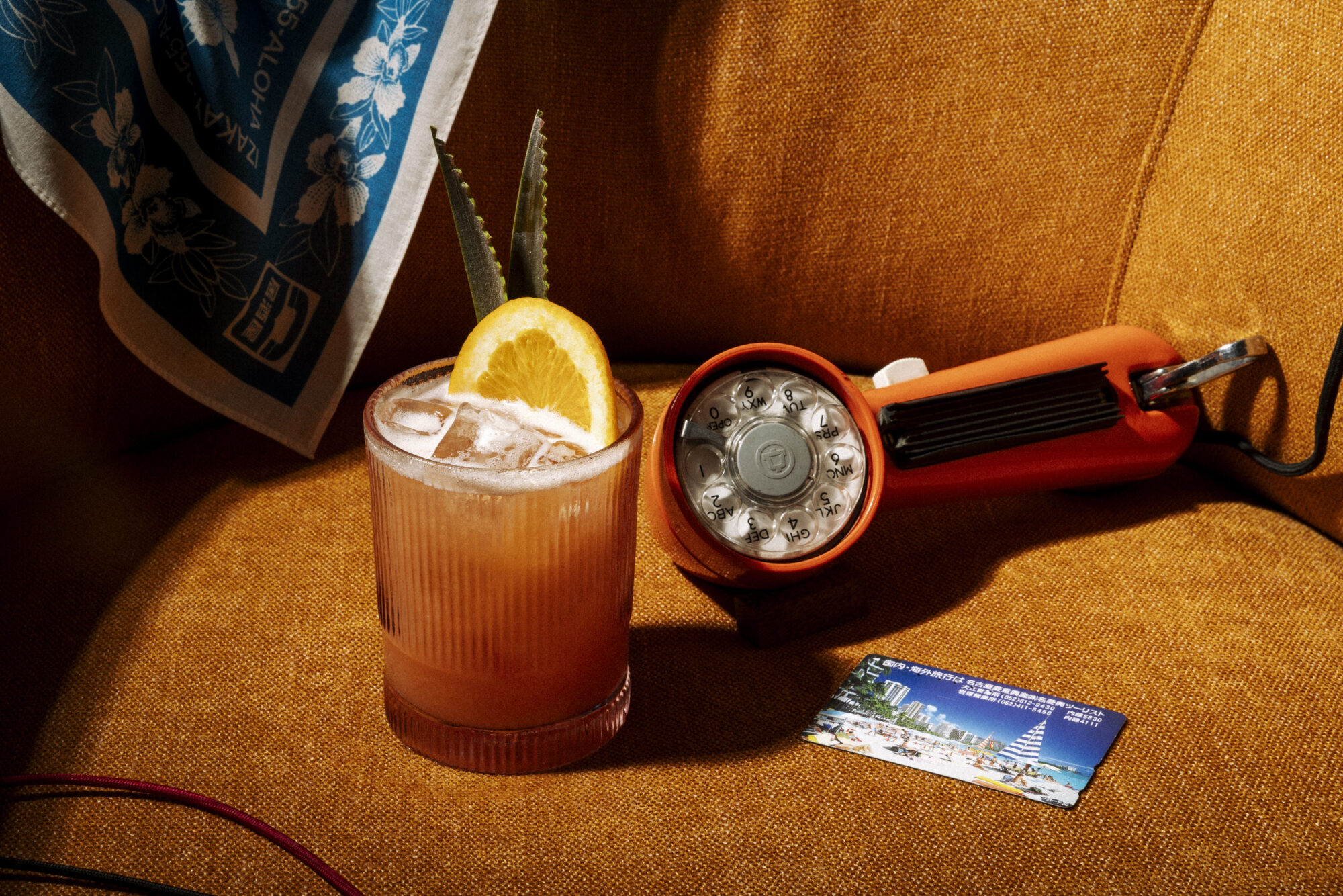 A cocktail with ice, an orange slice, and two leaves sits on an orange chair next to a vintage orange flashlight and a blue card with cityscape imagery. A blue-patterned cloth hangs in the background. captured by seattle's best food and hospitality photographer Brooke Fitts