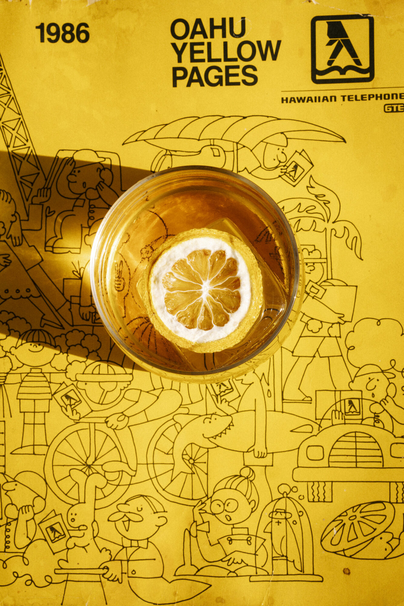 A glass of iced tea with a lemon slice sits on top of a bright yellow 1986 Oahu Yellow Pages phone book, featuring playful cartoon illustrations and bold black text. captured by seattle's best food and hospitality photographer Brooke Fitts