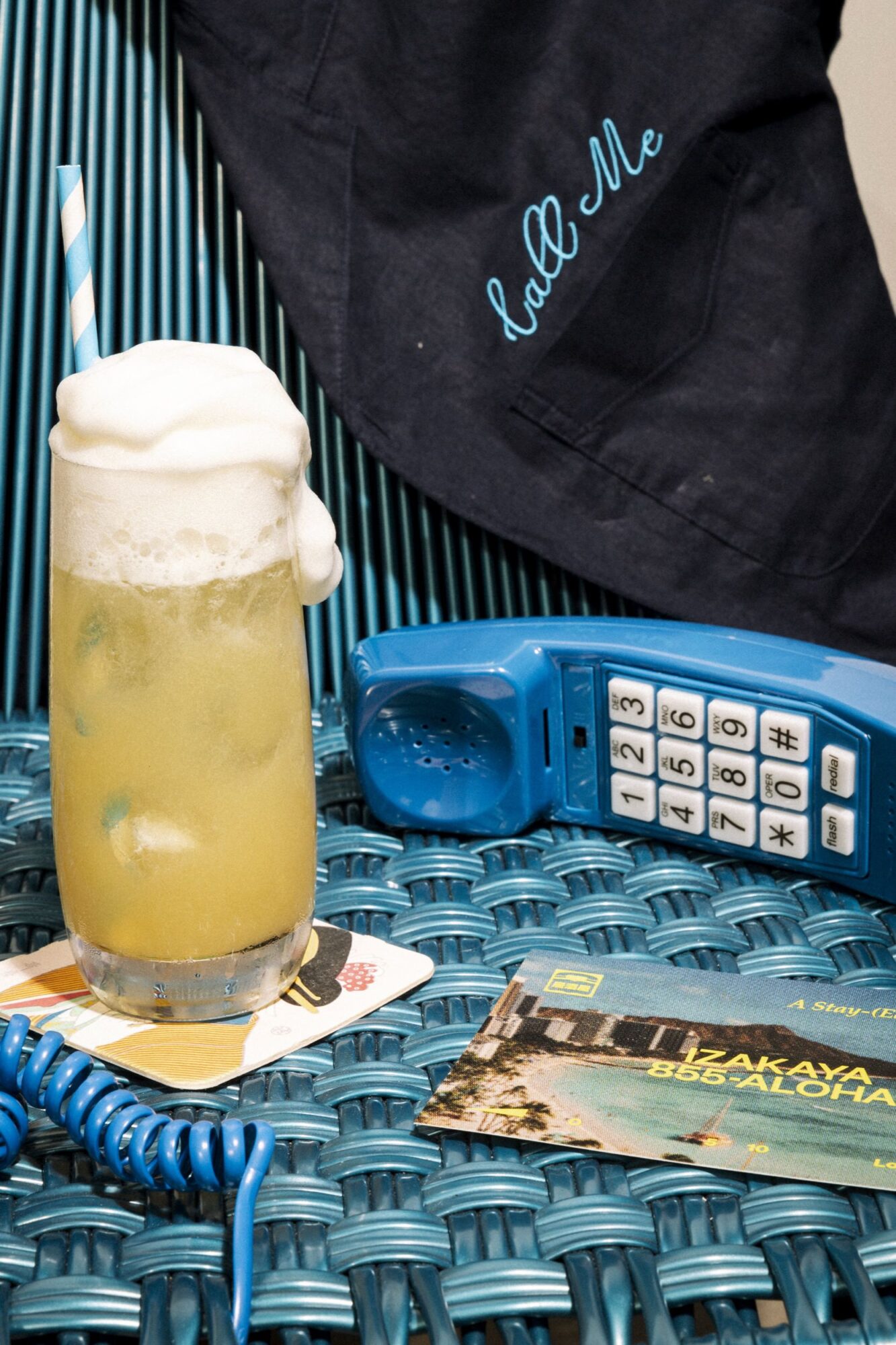 A creamy cocktail with a striped straw sits on a teal woven surface next to a blue retro phone, postcards, and a navy cloth with the words call me embroidered in blue. captured by seattle's best food and hospitality photographer Brooke Fitts