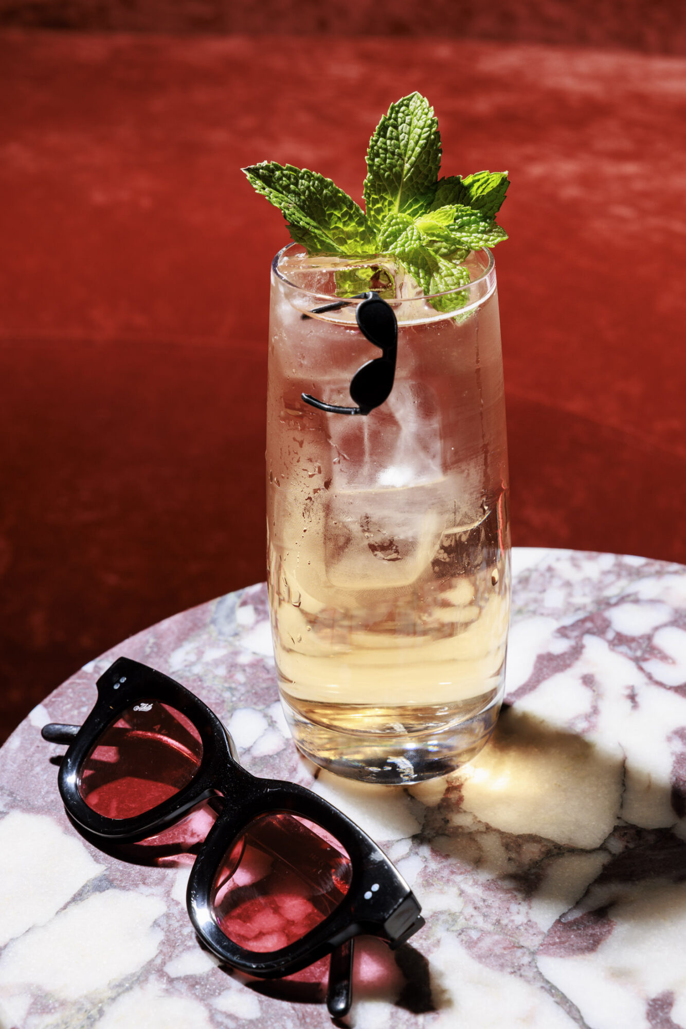 A tall glass with a light-colored drink, ice cubes, and a sprig of mint, sits on a marble table. A pair of black sunglasses lies nearby. The background is a rich red color. captured by seattle's best food and hospitality photographer Brooke Fitts