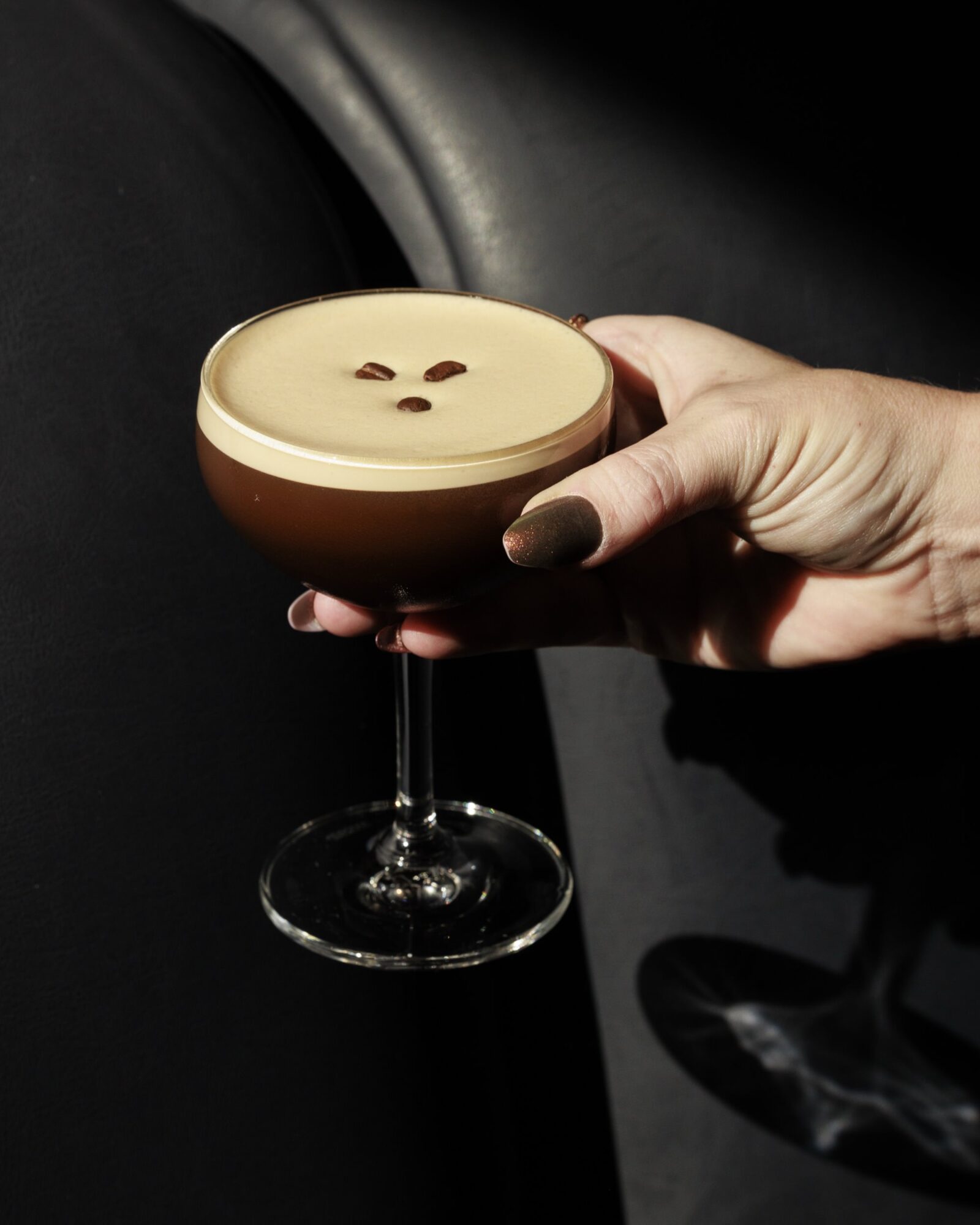 A hand with brown nail polish holds a coupe glass of espresso martini, topped with a creamy foam layer and three coffee beans as garnish, against a dark background. captured by seattle's best food and hospitality photographer Brooke Fitts