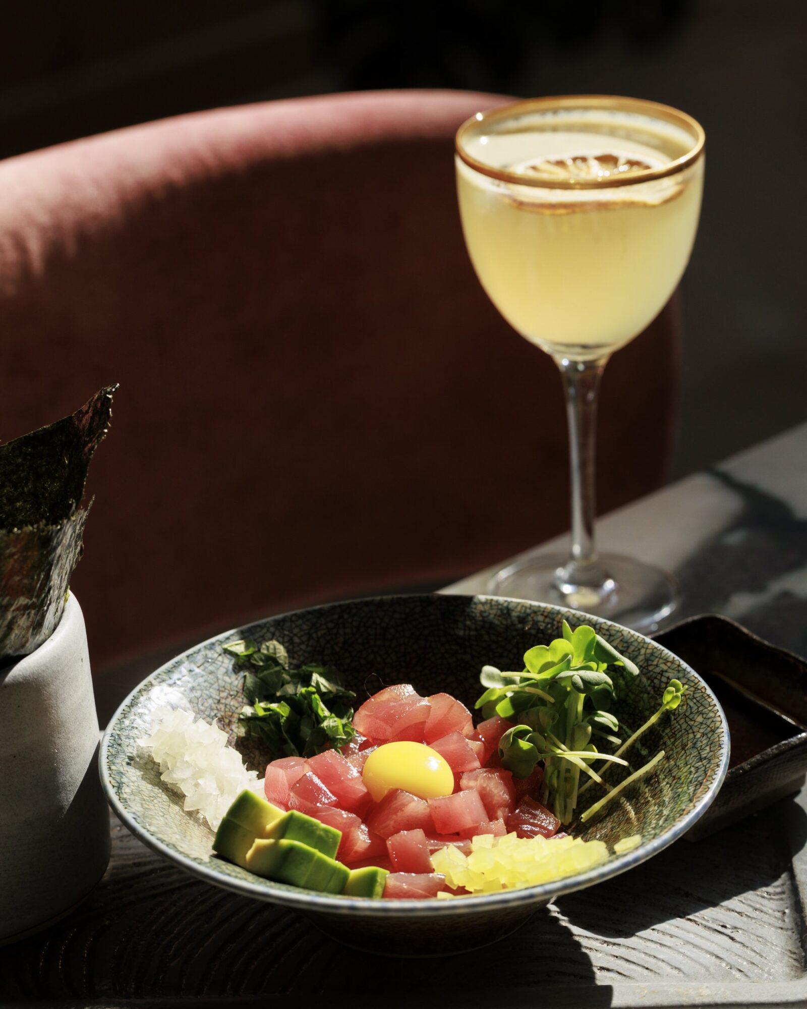 A bowl of poke with diced tuna, a raw egg yolk, cucumber, radish, and greens sits on a table next to a tall cocktail garnished with a dried citrus slice, all bathed in warm sunlight. captured by seattle's best food and hospitality photographer Brooke Fitts