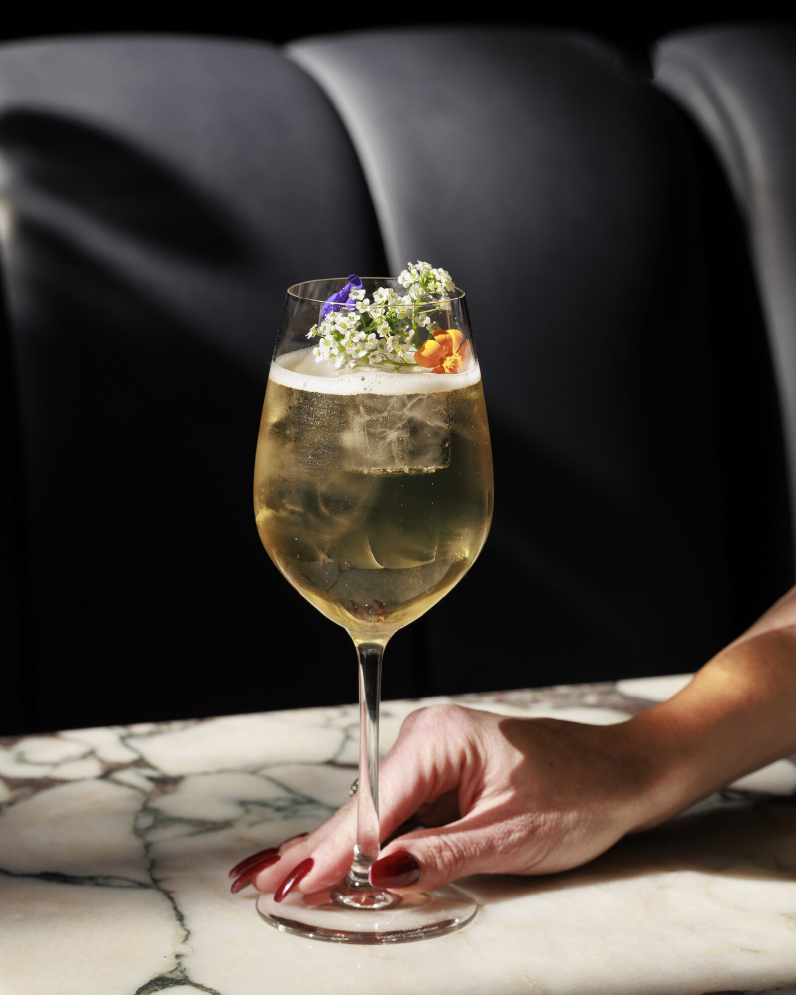 A hand with painted nails holds a wine glass filled with a light, iced cocktail garnished with colorful edible flowers and babys breath, resting on a marble table with a dark, blurred background. captured by seattle's best food and hospitality photographer Brooke Fitts