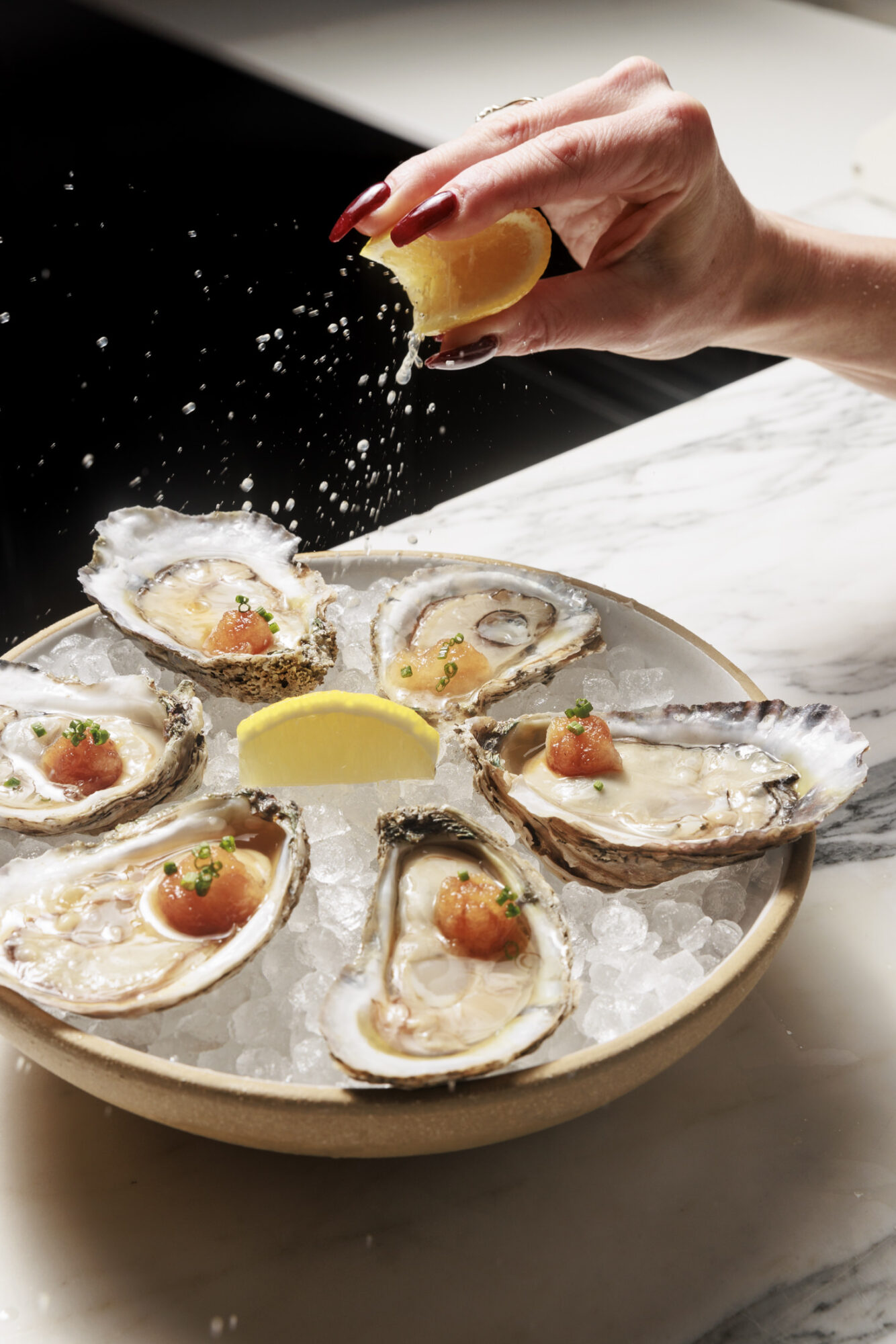 A hand squeezes a lemon wedge over a plate of fresh oysters on the half shell served on ice, each topped with a small garnish. captured by seattle's best food and hospitality photographer Brooke Fitts