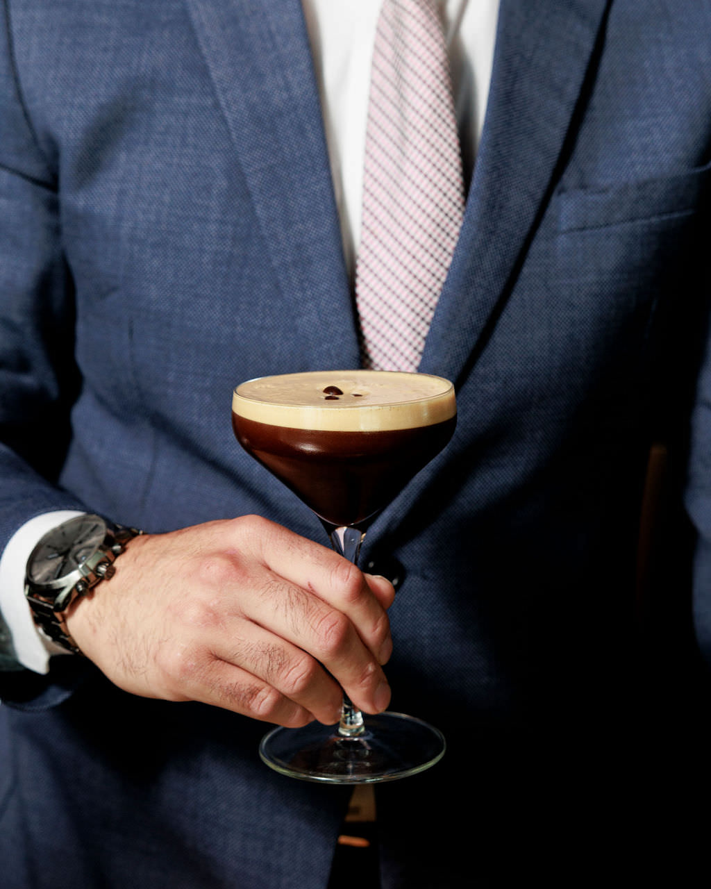 A person in a blue suit and light pink patterned tie holds a stemmed glass with a dark espresso martini topped with foam and coffee beans. captured by seattle's best food and hospitality photographer Brooke Fitts