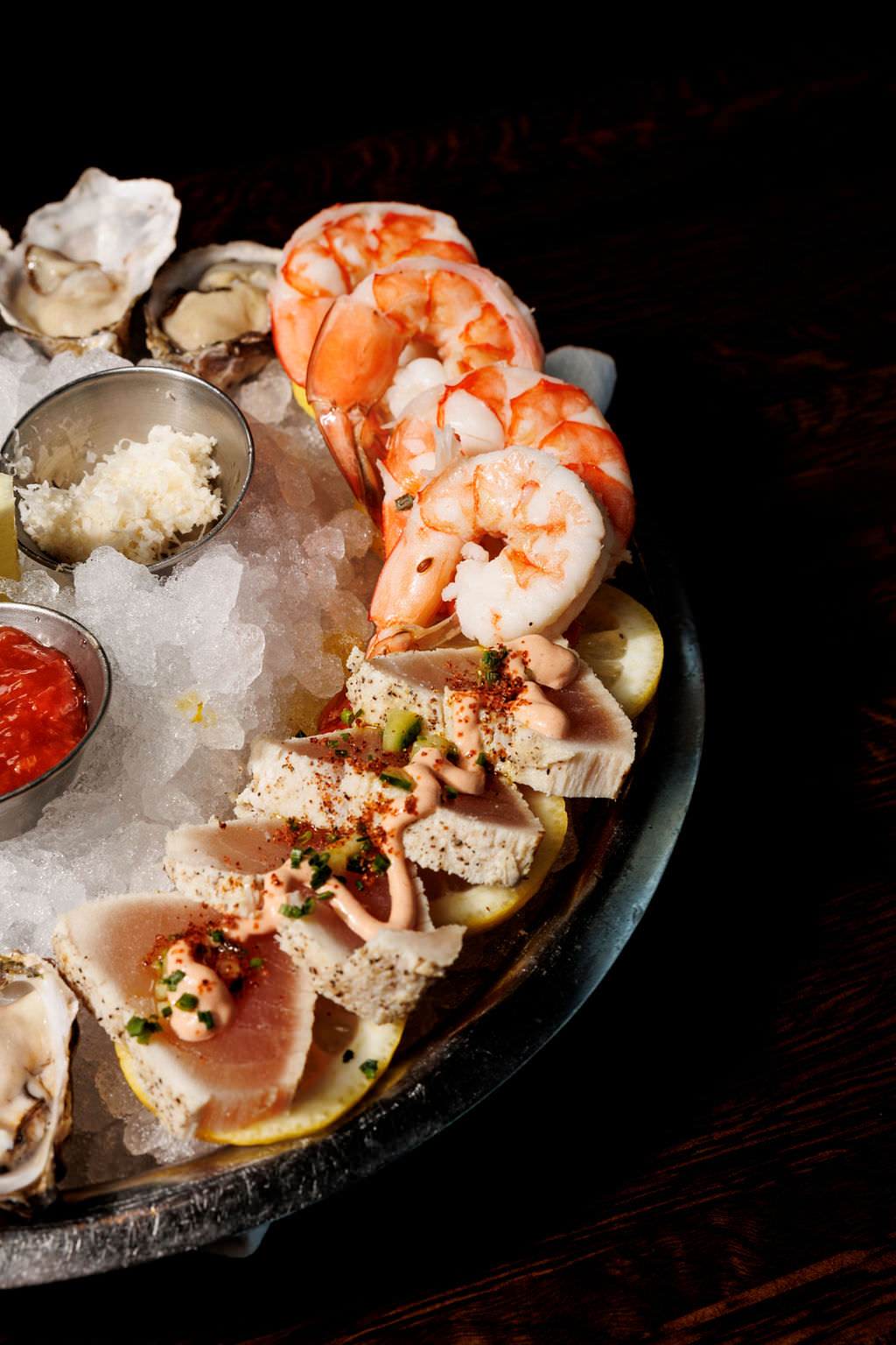 A seafood platter on ice featuring shrimp, oysters, and slices of seared tuna, served with lemon wedges and dipping sauces, on a dark wooden surface. captured by seattle's best food and hospitality photographer Brooke Fitts