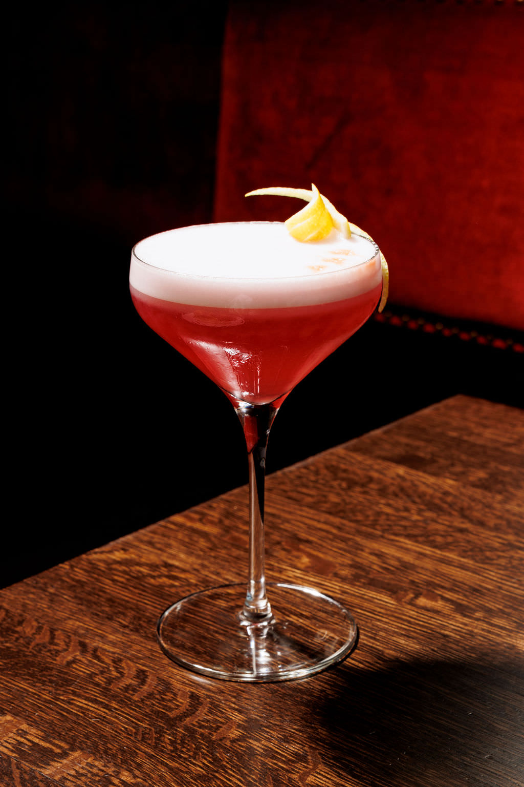 A pink cocktail with a foamy top is served in a stemmed glass, garnished with a twist of lemon peel. The drink sits on a dark wooden table with a red upholstered background. captured by seattle's best food and hospitality photographer Brooke Fitts