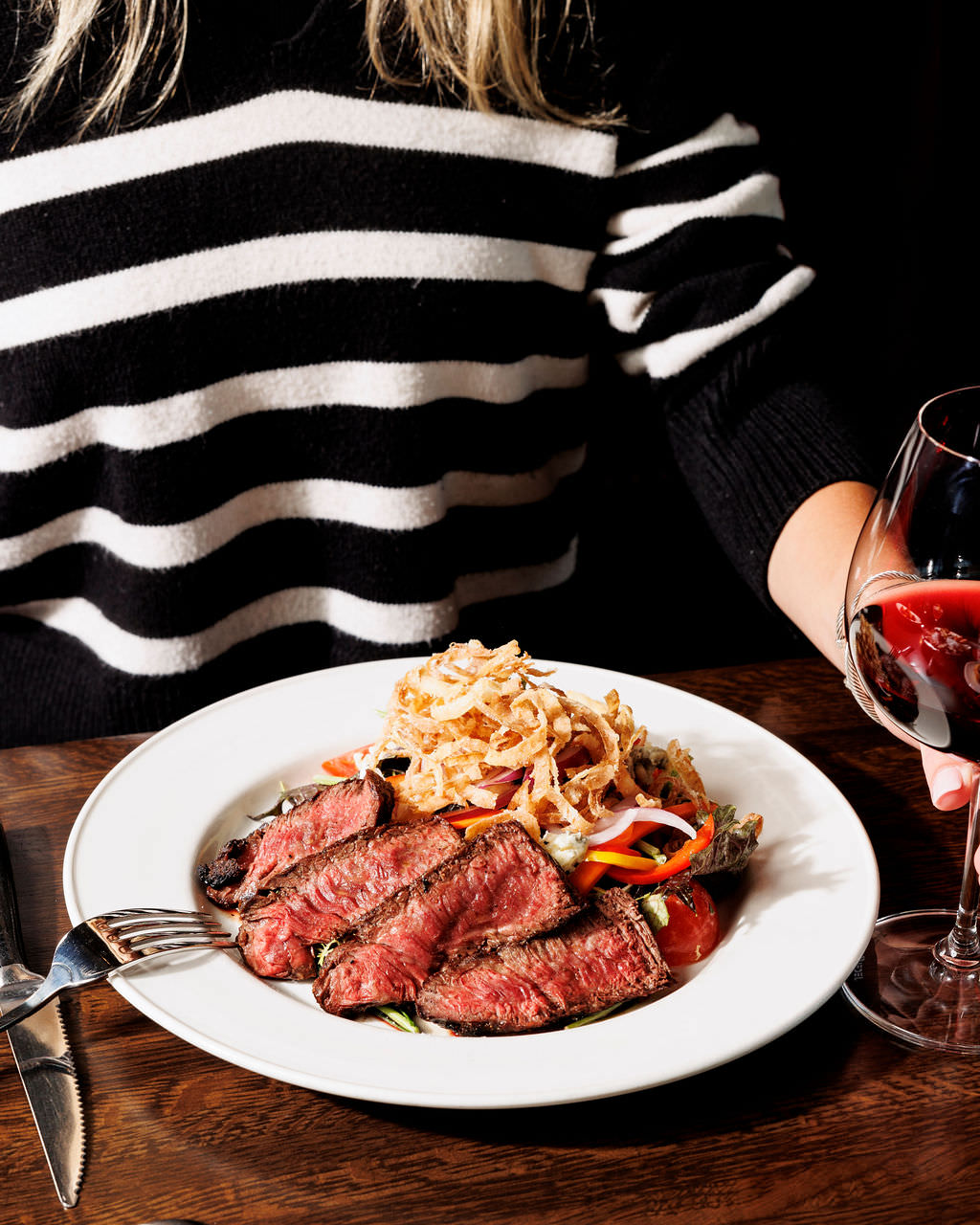 A person in a black and white striped sweater sits at a table with a plate of sliced steak, crispy onions, and vegetables, holding a glass of red wine. captured by seattle's best food and hospitality photographer Brooke Fitts