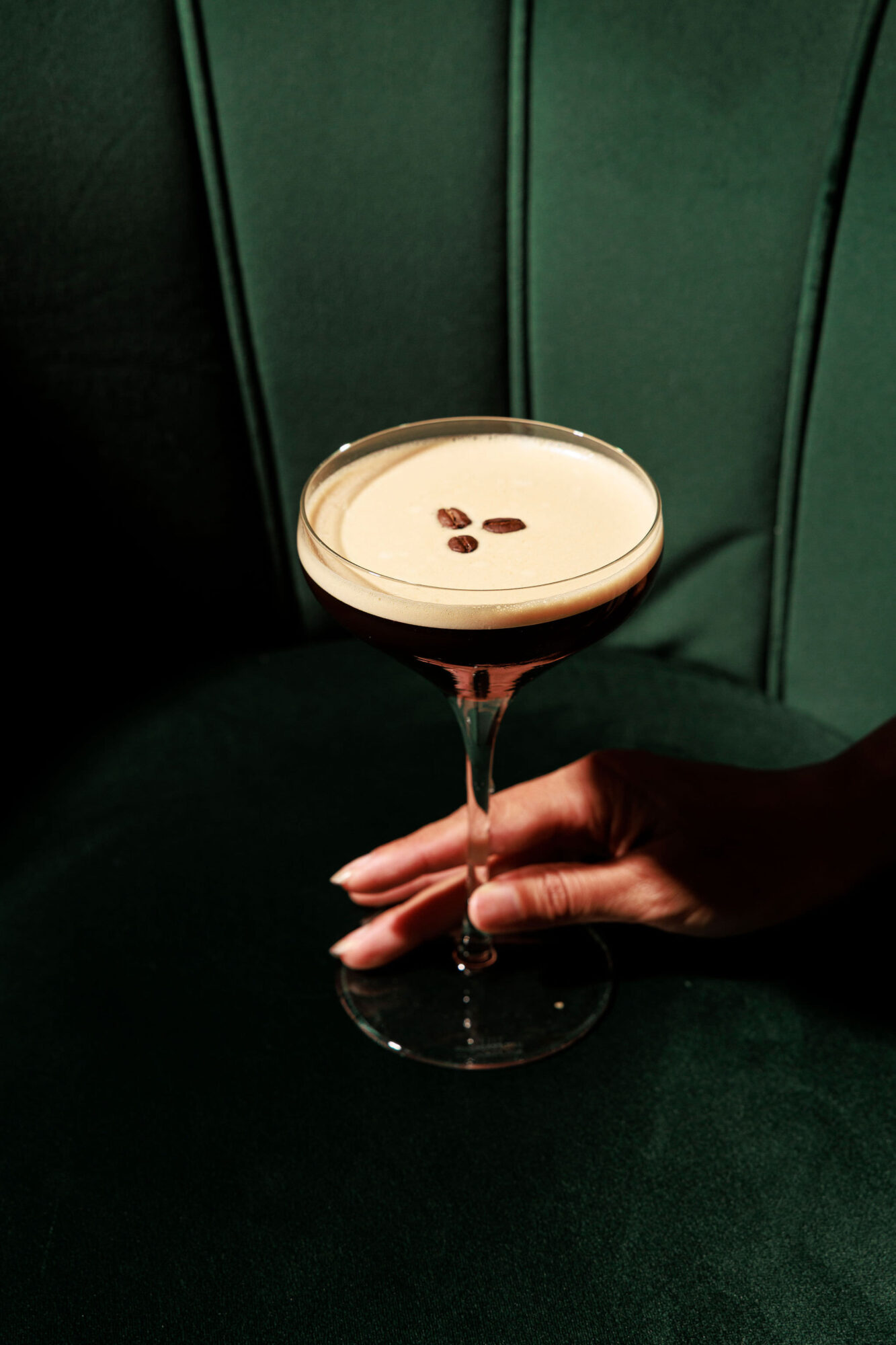 A hand holds a coupe glass filled with an espresso martini, topped with three coffee beans, against a dark green velvet background. captured by seattle's best food and hospitality photographer Brooke Fitts