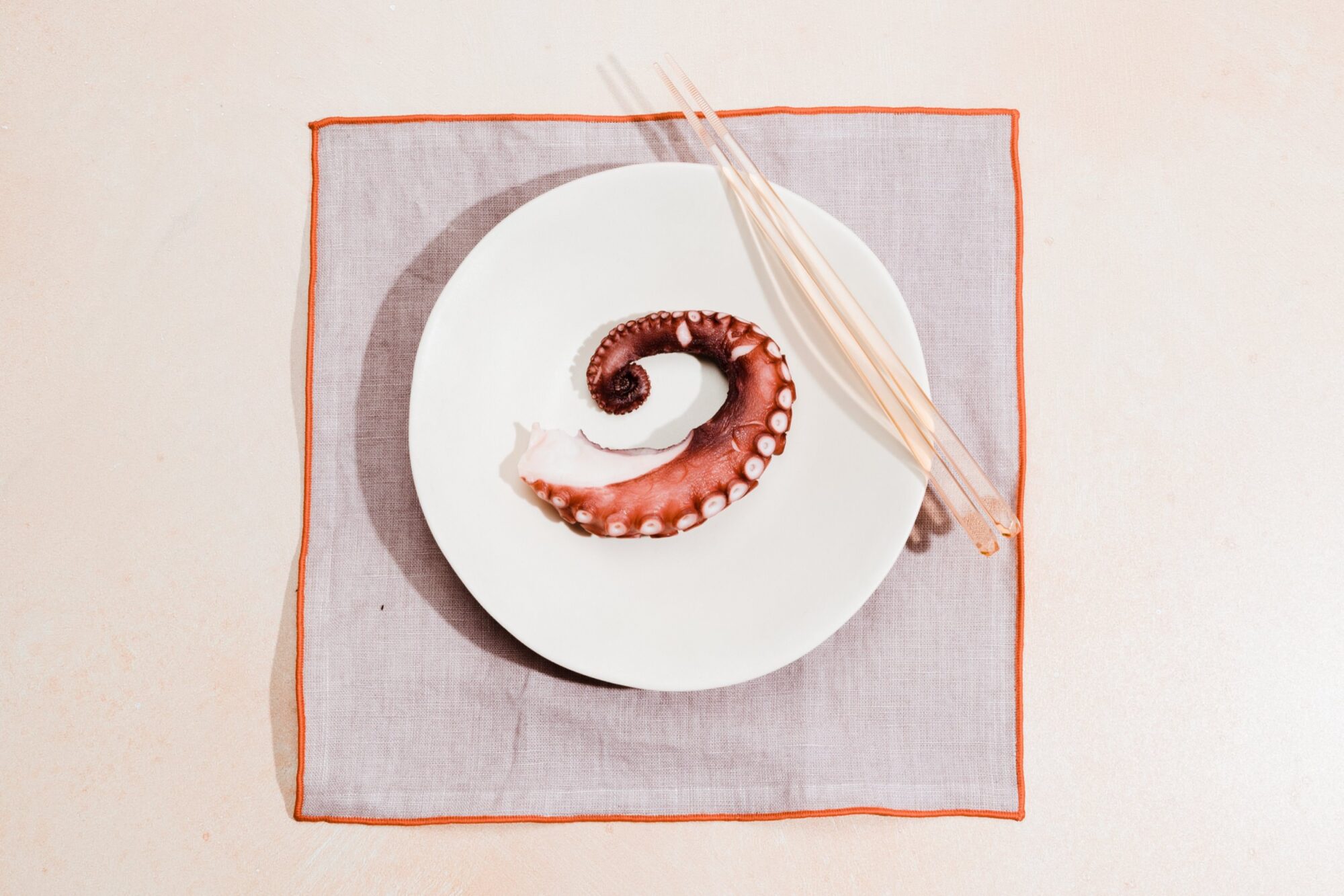 A single octopus tentacle on a white plate, placed on a gray napkin with an orange border, with a pair of wooden chopsticks resting on the plate, all set on a light surface. captured by seattle's best food and hospitality photographer Brooke Fitts