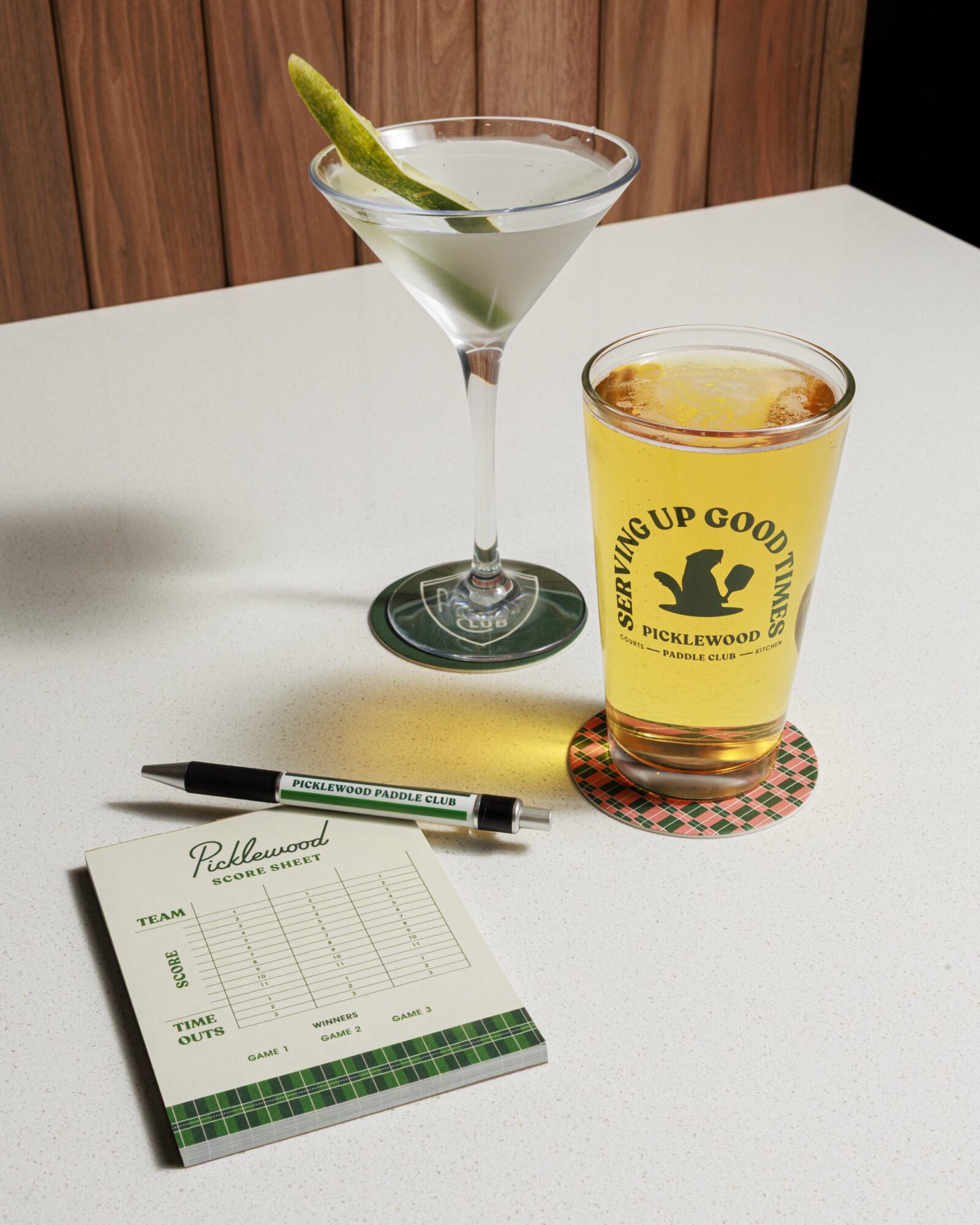 A martini with a pickle garnish, a pint glass of beer with Serving Up Good Times Picklewood Paddle Club text, a pen, and a Picklewood score sheet sit on a white countertop. captured by seattle's best food and hospitality photographer Brooke Fitts