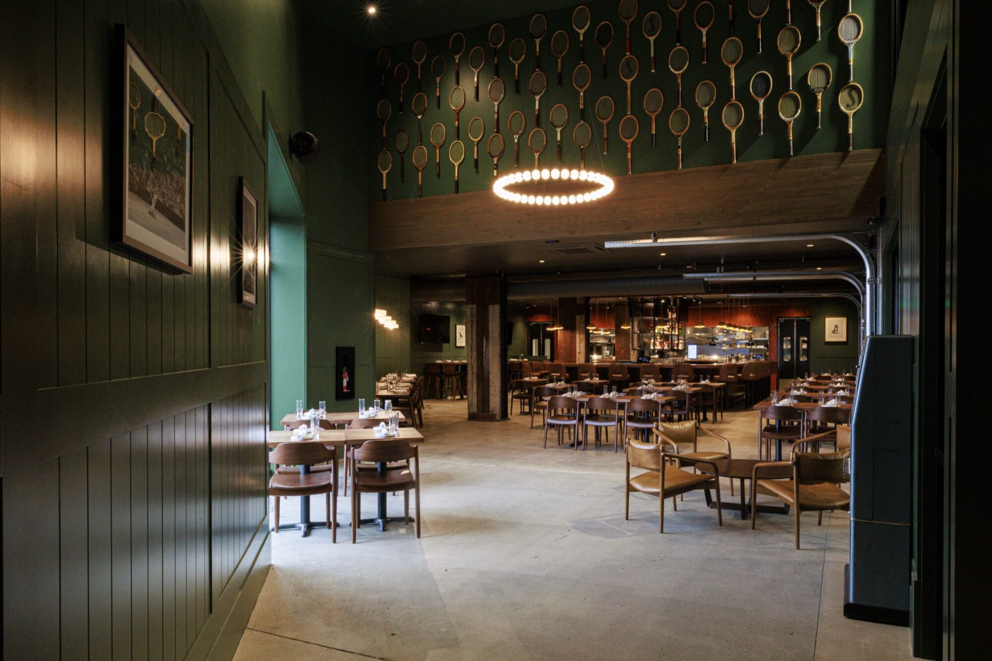 A stylish restaurant with green walls, wooden tables and chairs, and a modern circular chandelier. The upper wall is decorated with many tennis rackets, and the open kitchen and bar are visible in the background. captured by seattle's best food and hospitality photographer Brooke Fitts