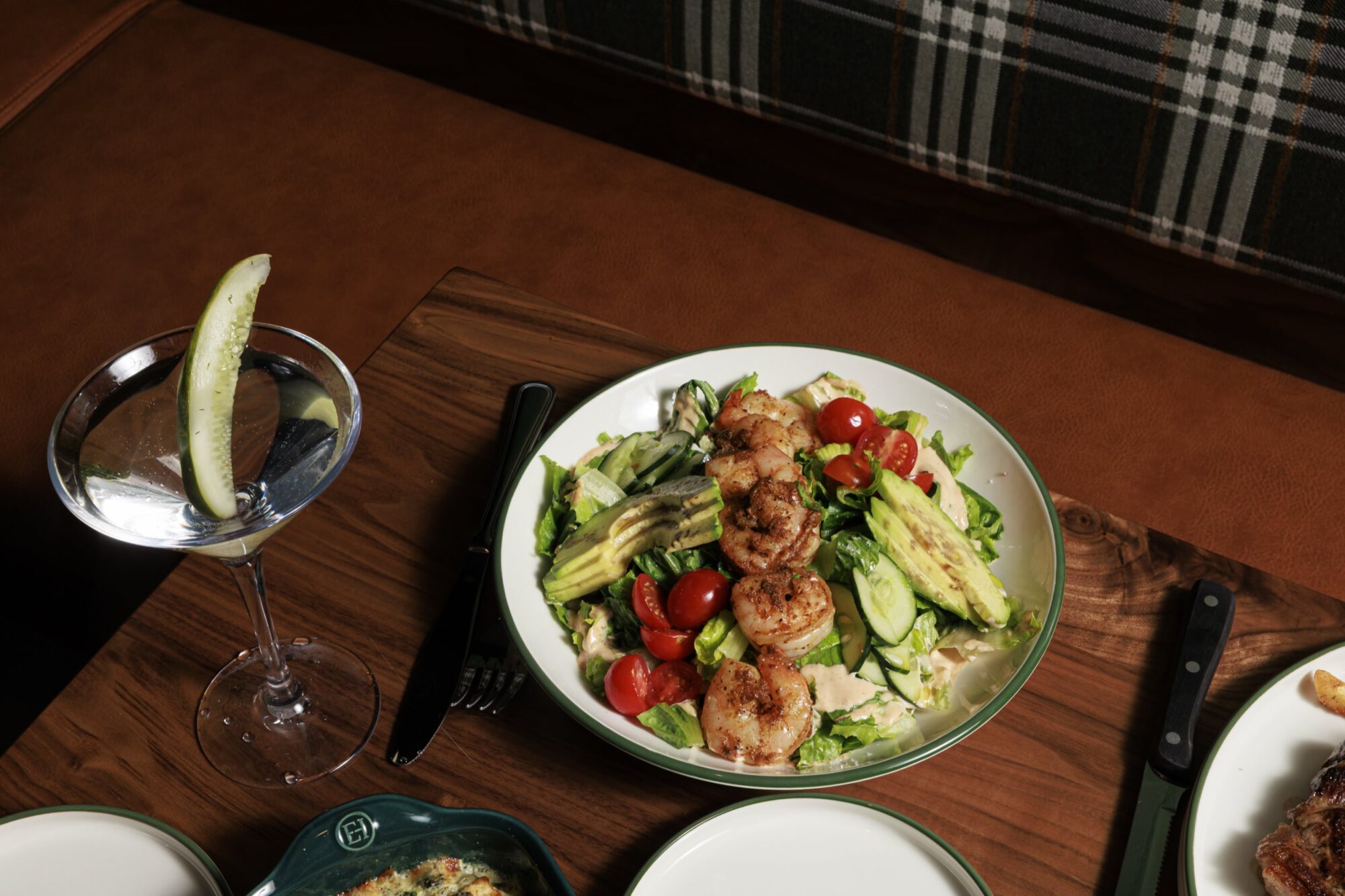 A fresh salad with grilled shrimp, avocado, cucumber, and cherry tomatoes sits on a wooden table next to a cocktail garnished with a pickle spear. A knife and fork are placed beside the plate. captured by seattle's best food and hospitality photographer Brooke Fitts