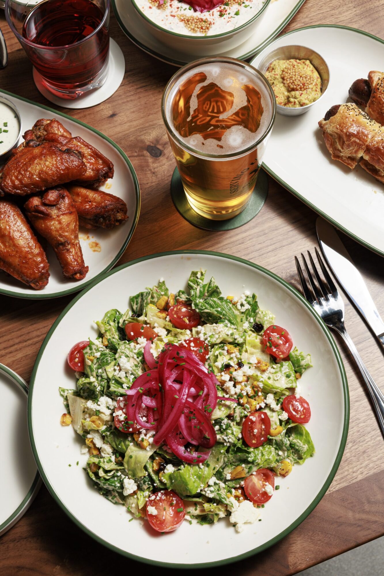 A table set with a green salad topped with cherry tomatoes, feta, and pickled onions, alongside chicken wings, pigs in a blanket, a glass of beer, a red drink, and creamy dip with bread. captured by seattle's best food and hospitality photographer Brooke Fitts