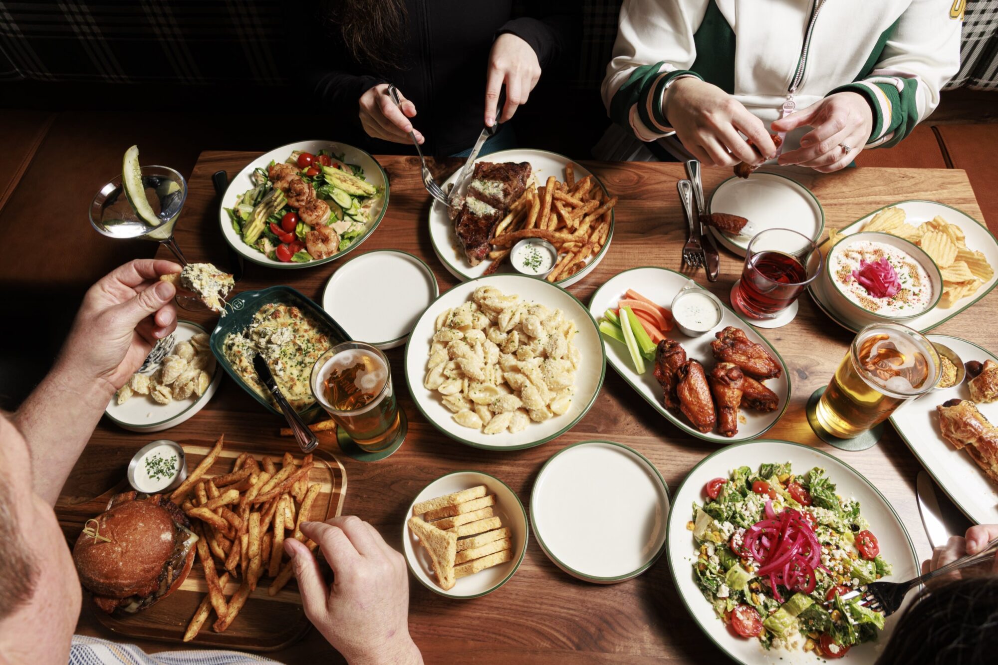 Four people dine at a table filled with various dishes, including fries, pasta, salads, chicken wings, a burger, bread, vegetables with dip, drinks, and sauces. Hands are reaching for and serving the food. captured by seattle's best food and hospitality photographer Brooke Fitts
