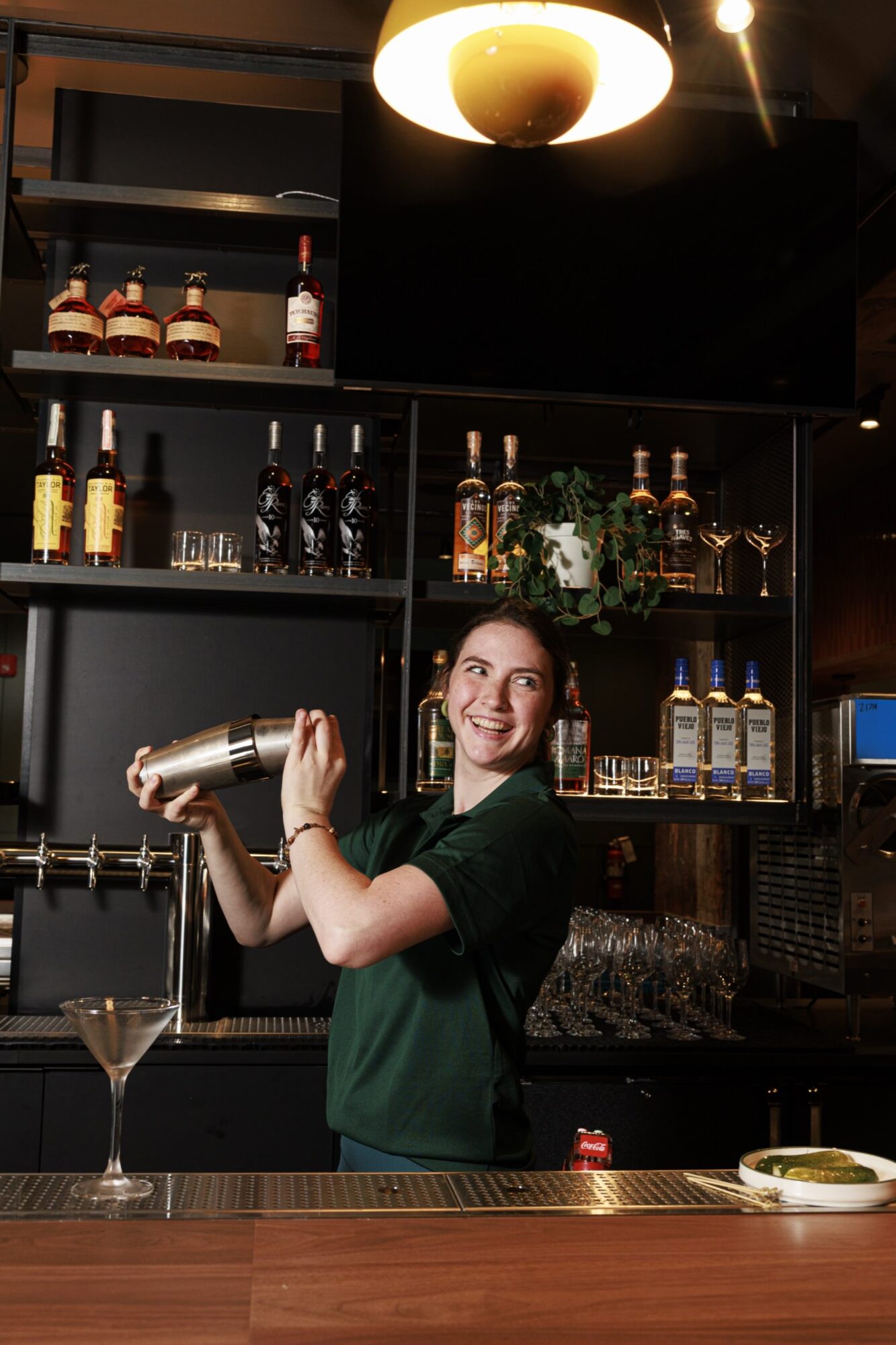 A smiling bartender in a green shirt shakes a cocktail shaker behind a modern bar, with bottles and glasses on shelves, a martini glass and a plate of olives on the counter in front of her. captured by seattle's best food and hospitality photographer Brooke Fitts