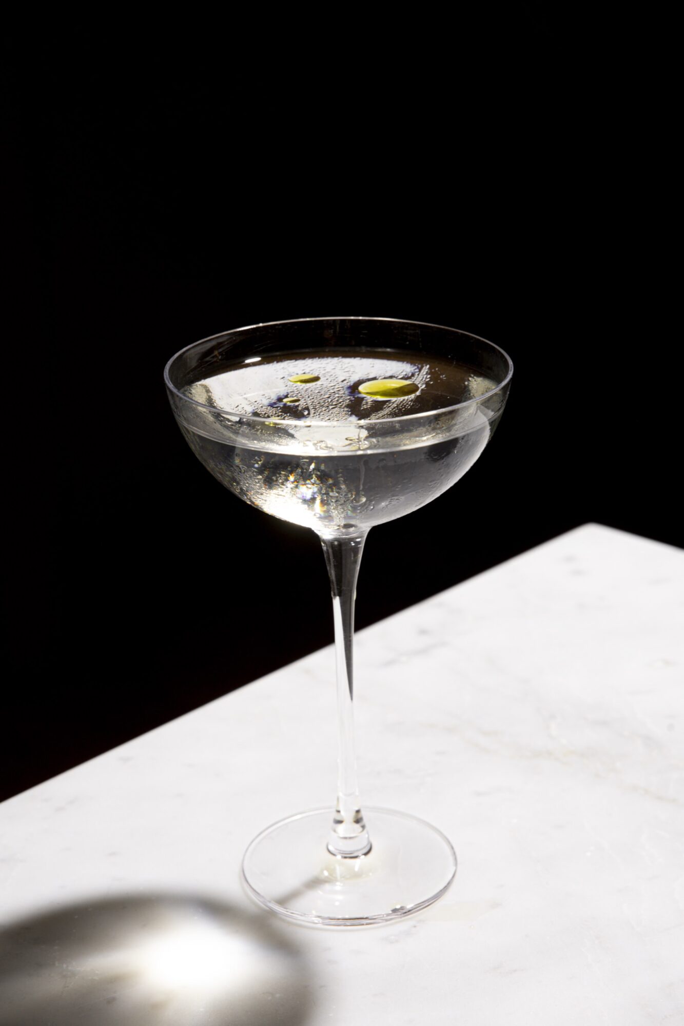 A clear martini glass filled with a transparent cocktail, garnished with a few green olives, sits on a white marble surface against a black background. captured by seattle's best food and hospitality photographer Brooke Fitts