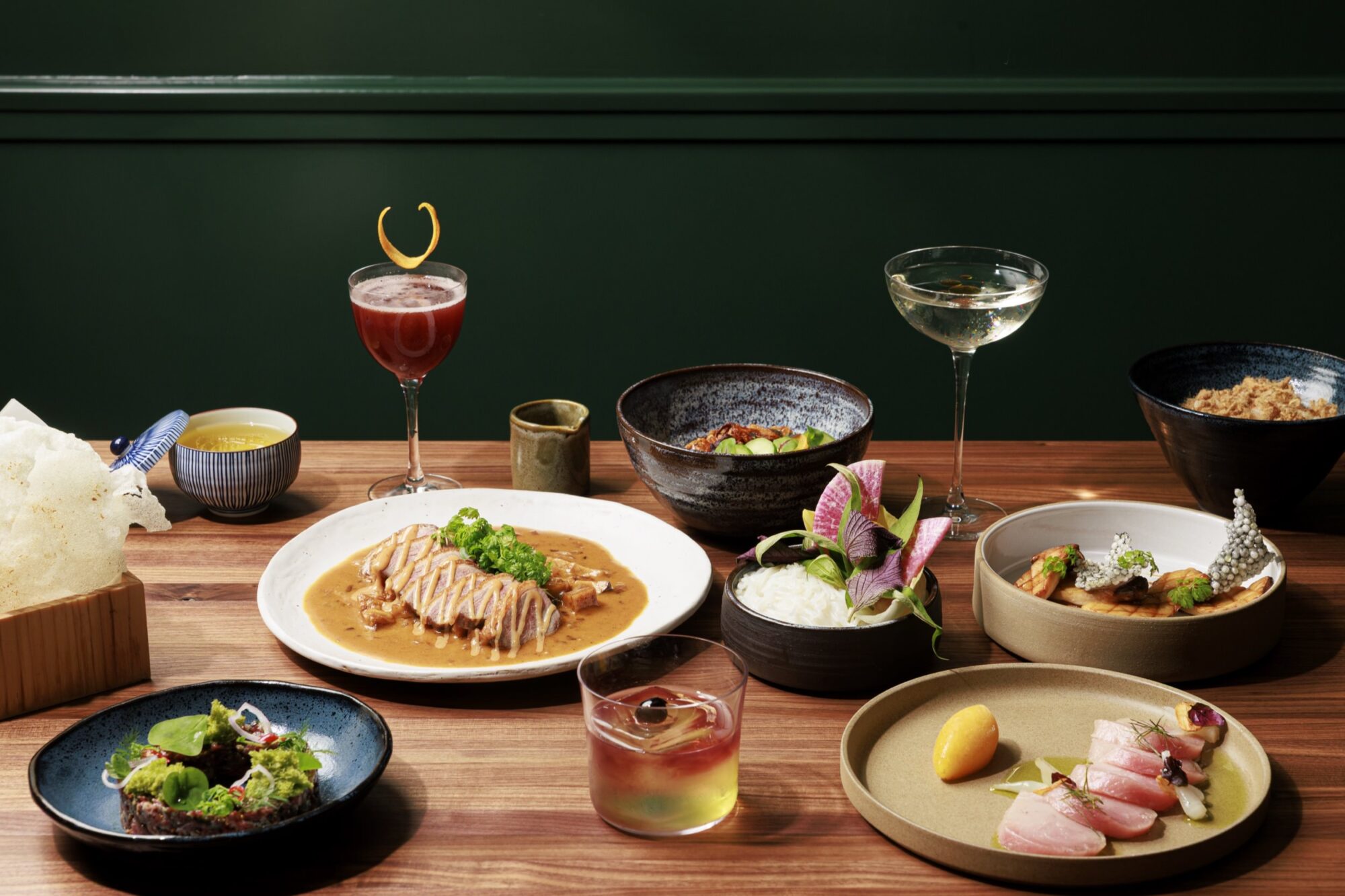 A beautifully arranged table features various gourmet dishes, cocktails, and bowls with rice, salads, and sauces against a dark green wall, suggesting an elegant fine dining experience. captured by seattle's best food and hospitality photographer Brooke Fitts