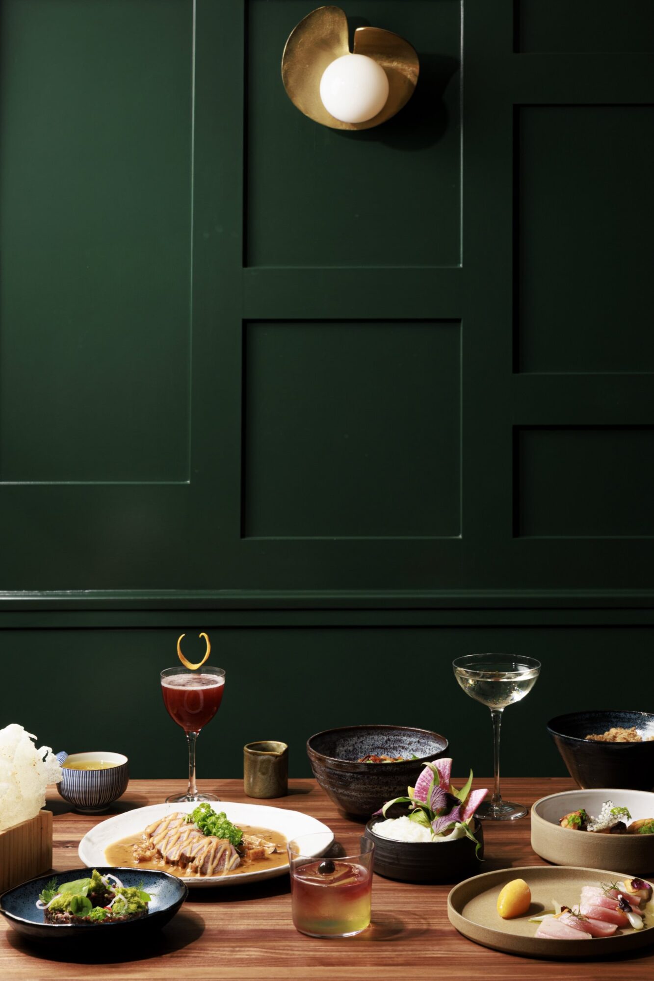 A wooden table set with assorted gourmet dishes and cocktails, placed against a dark green paneled wall with a modern brass and white wall sconce overhead. captured by seattle's best food and hospitality photographer Brooke Fitts